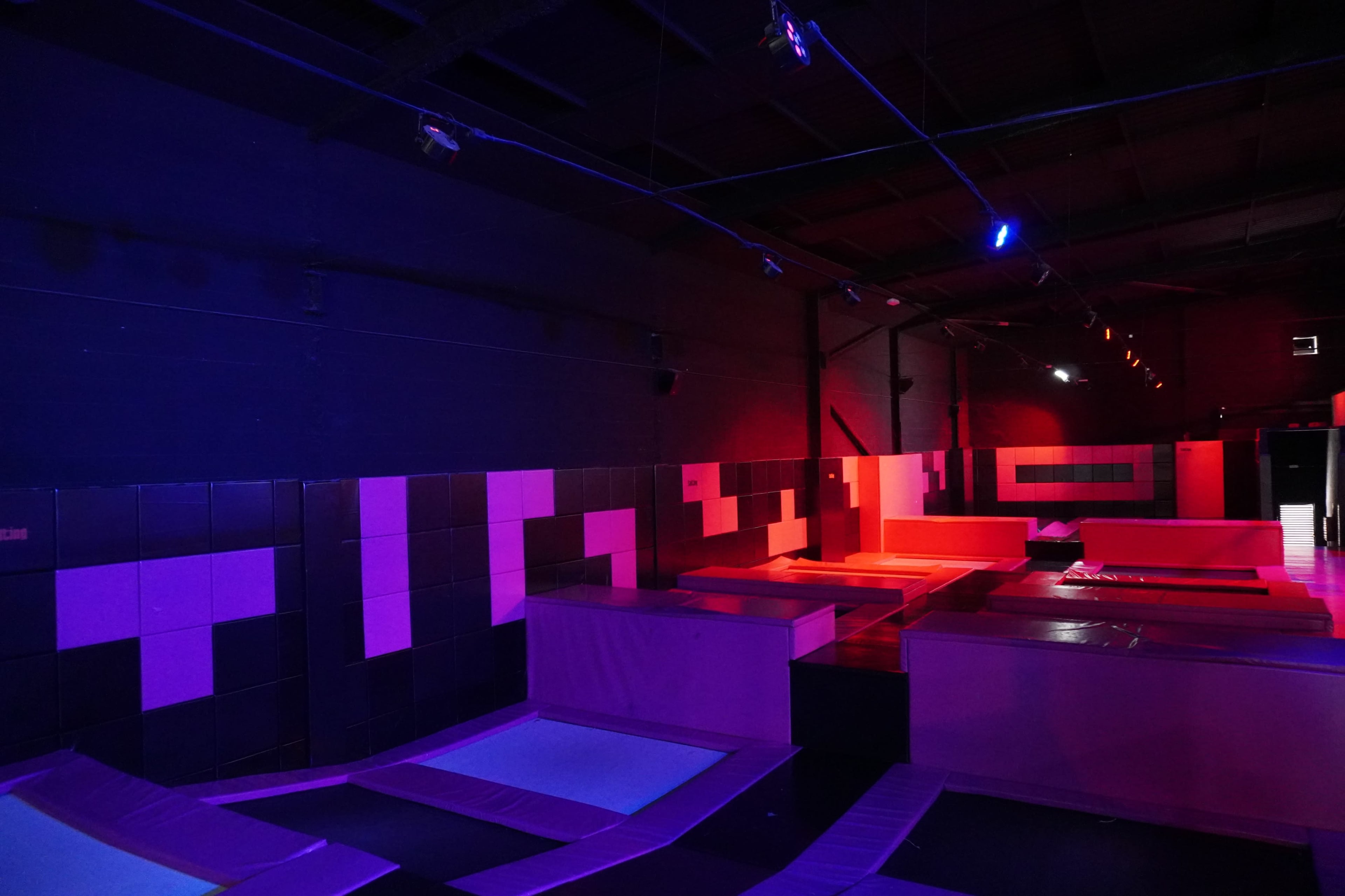 An indoor trampoline park with various blue and red trampolines arranged in a dimly lit space featuring colorful lighting.