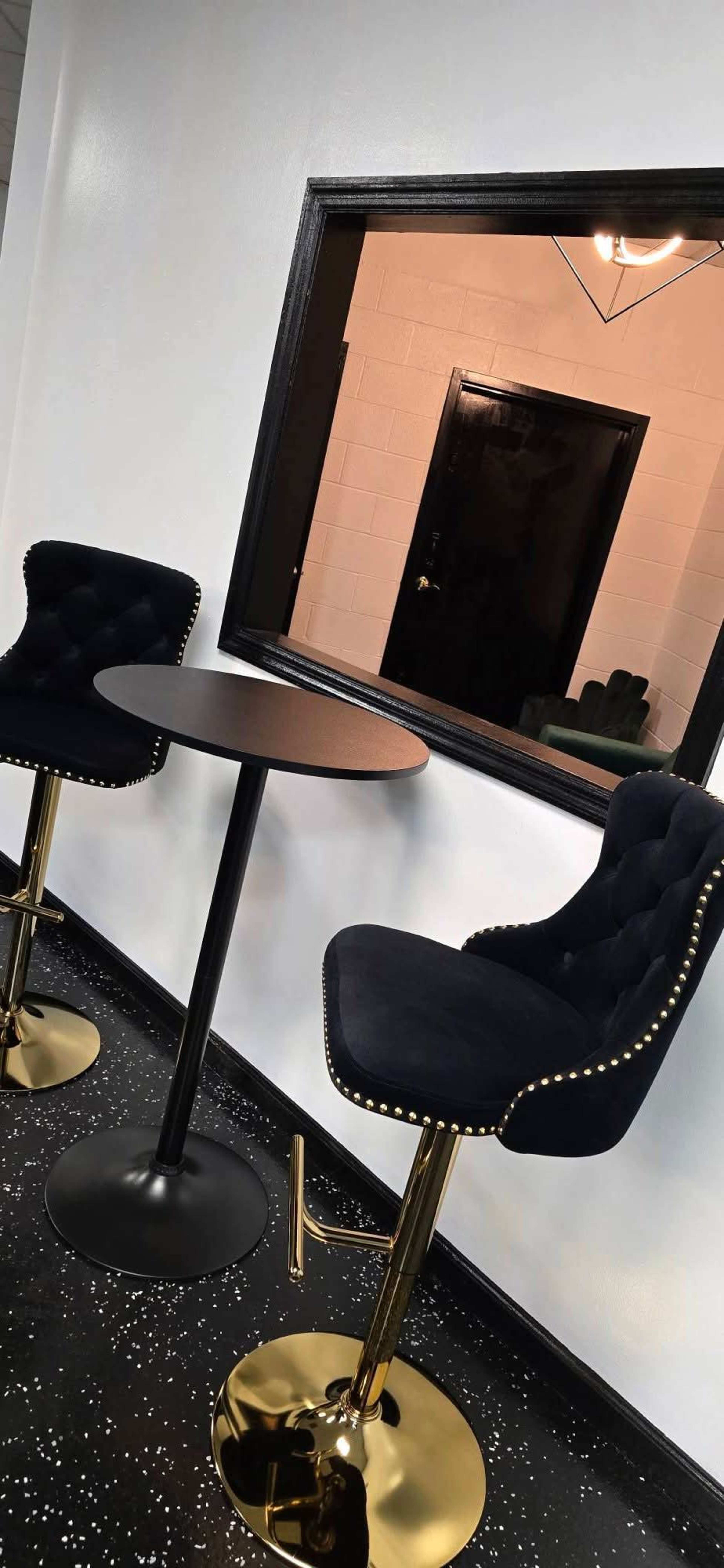 The image shows two black upholstered bar stools with gold accents positioned next to a round table, against a white wall featuring a rectangular mirror reflecting a door.