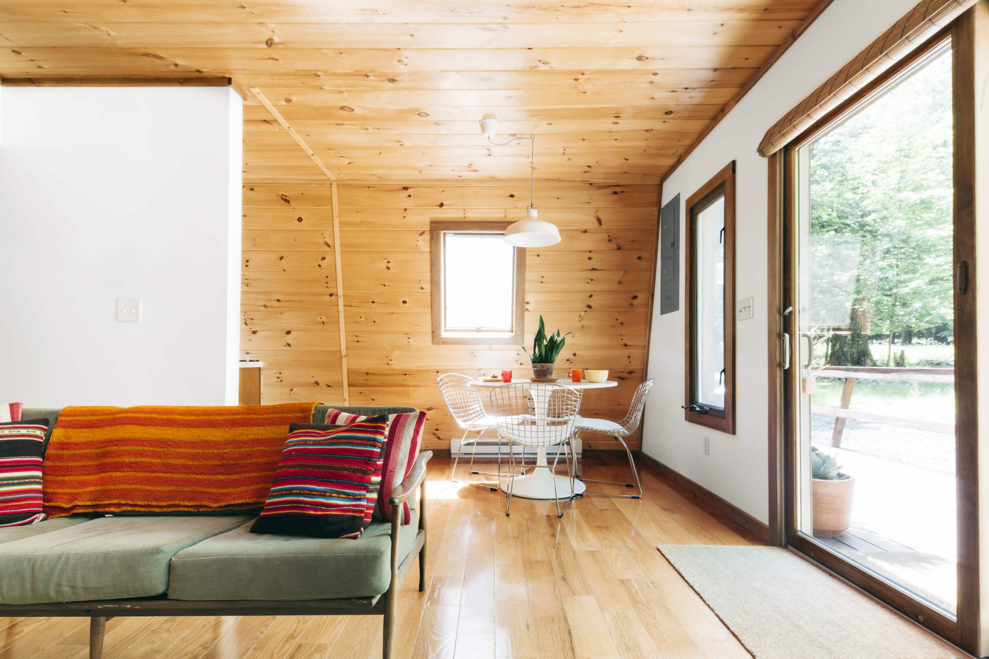 A wooden-paneled living area with a green sofa, colorful pillows, a round dining table, and a large window overlooking an outdoor view.