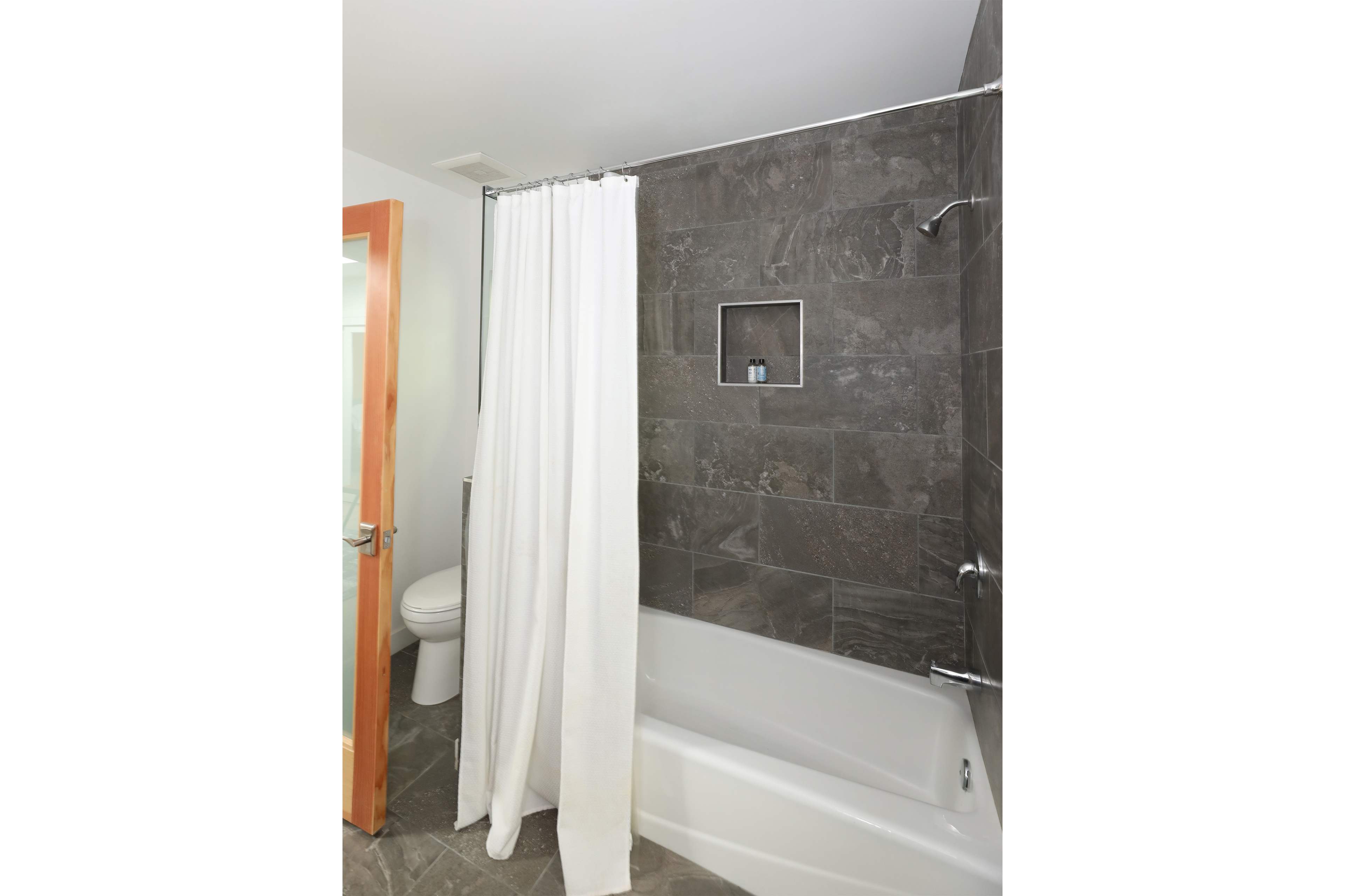 A bathroom features a bathtub with a shower curtain, tiled walls, and a toilet in the background.