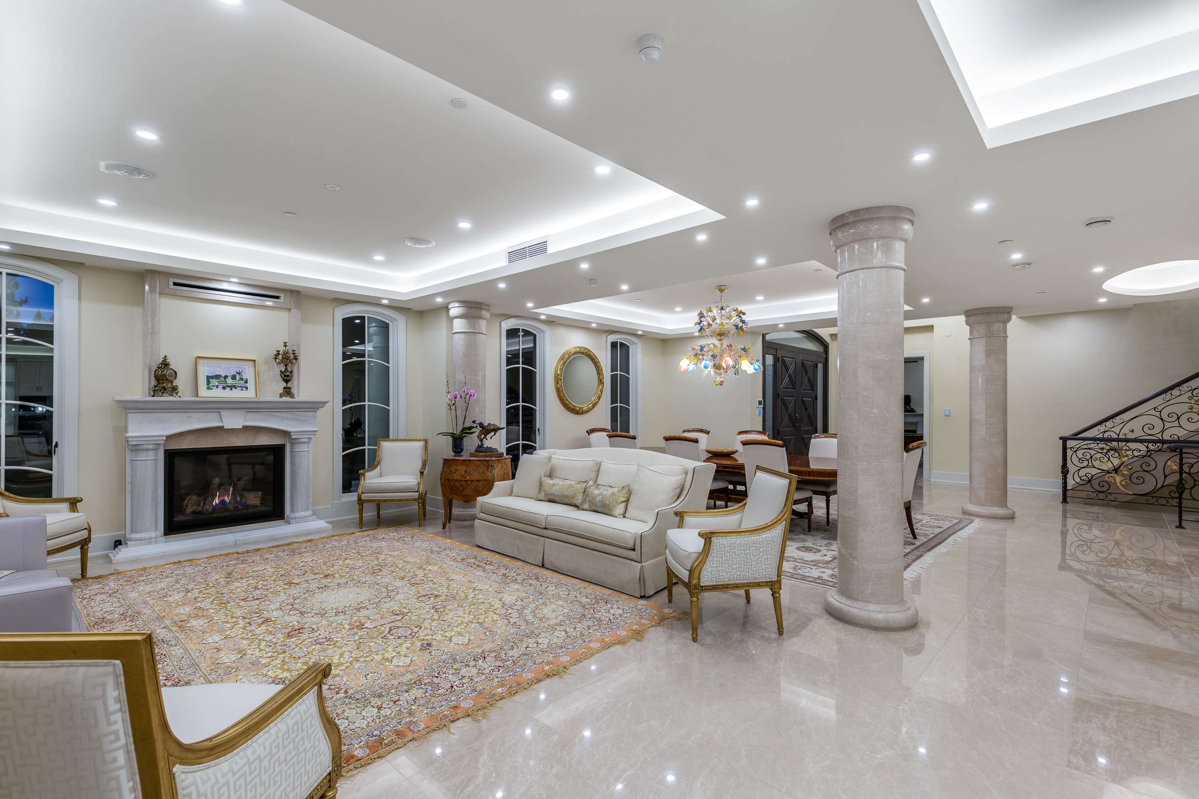 The image shows a luxurious living room featuring a fireplace, elegant seating, and a chandelier, with marble flooring and decorative pillars.
