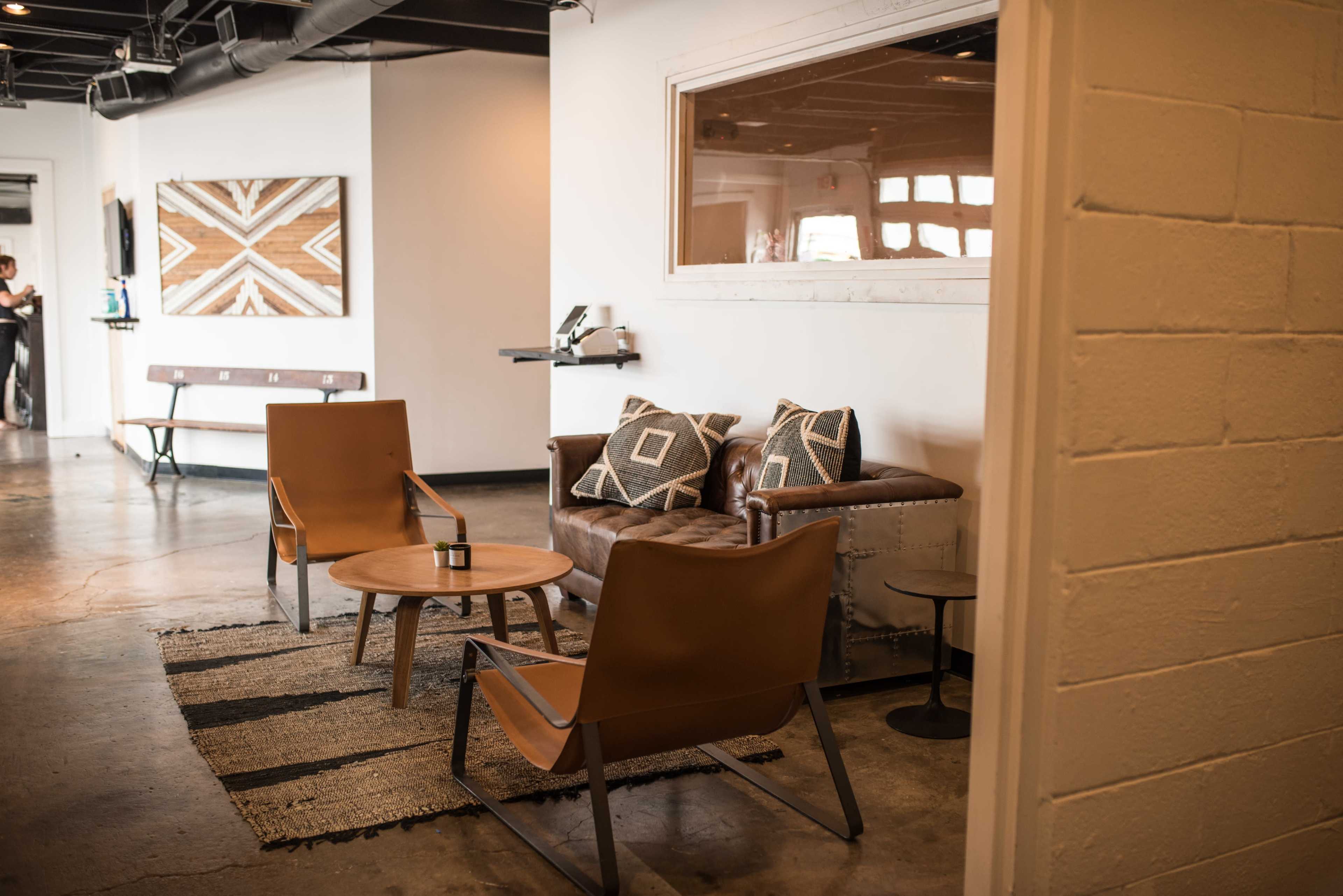 A modern lounge area with two brown chairs, a brown leather couch, a small round table, and a patterned rug on a concrete floor.