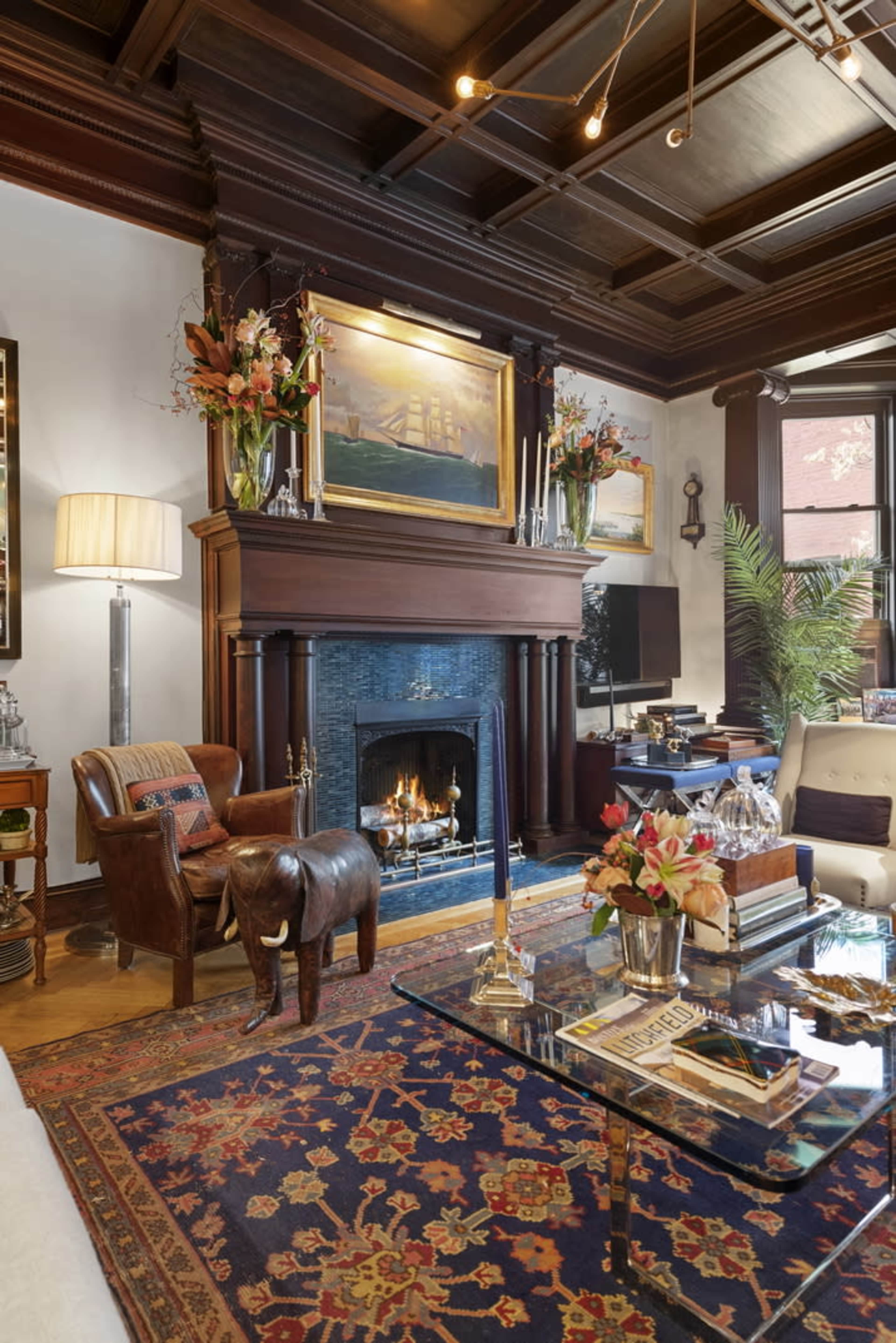 The scene features a cozy living room with a dark wood-beamed ceiling, a fireplace framed by a decorative mantel, and elegant furnishings including a leather armchair and a glass coffee table.