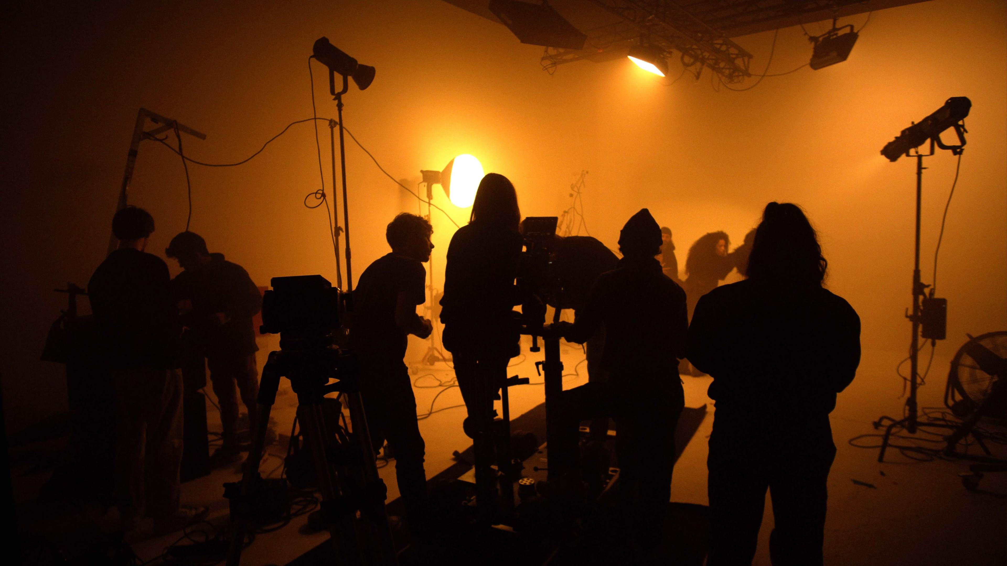 A group of silhouetted individuals operates film equipment in a dimly lit studio with warm lighting.