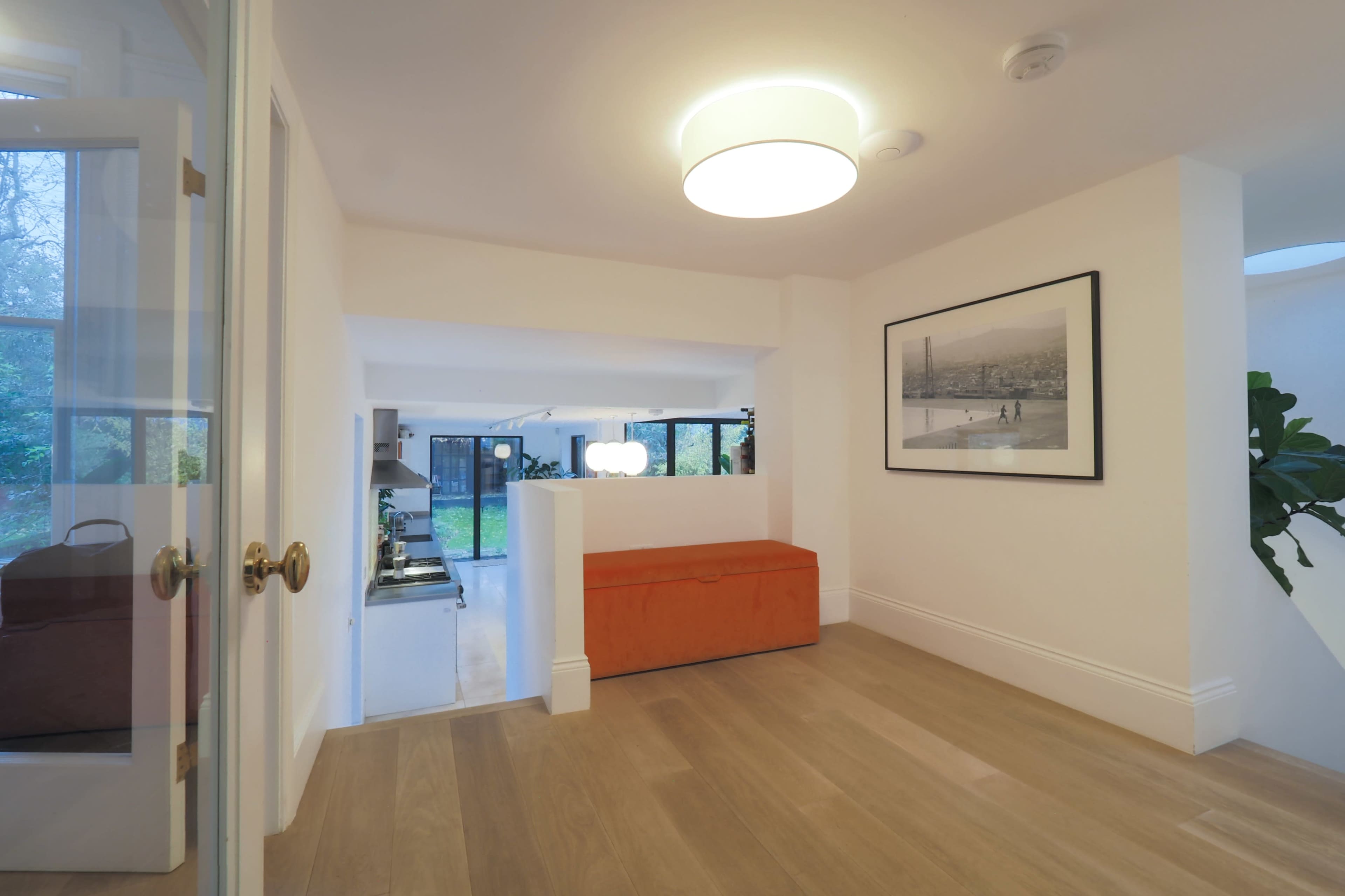 The image shows a modern interior hallway with a light fixture, an orange bench, and a doorway leading to a spacious kitchen area.