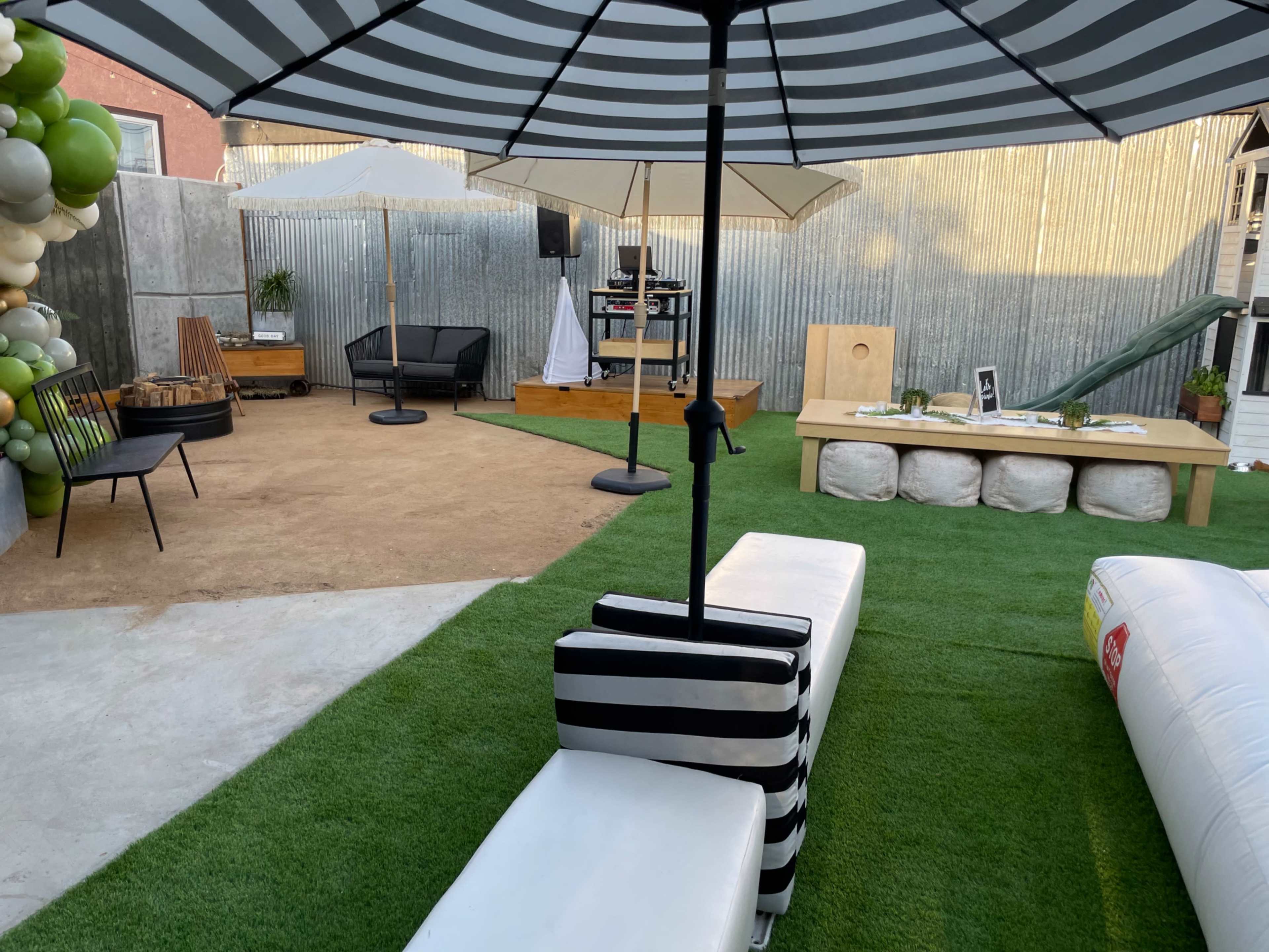A landscaped outdoor area featuring seating, an umbrella, decorative elements, and a stage setup against a corrugated metal backdrop.