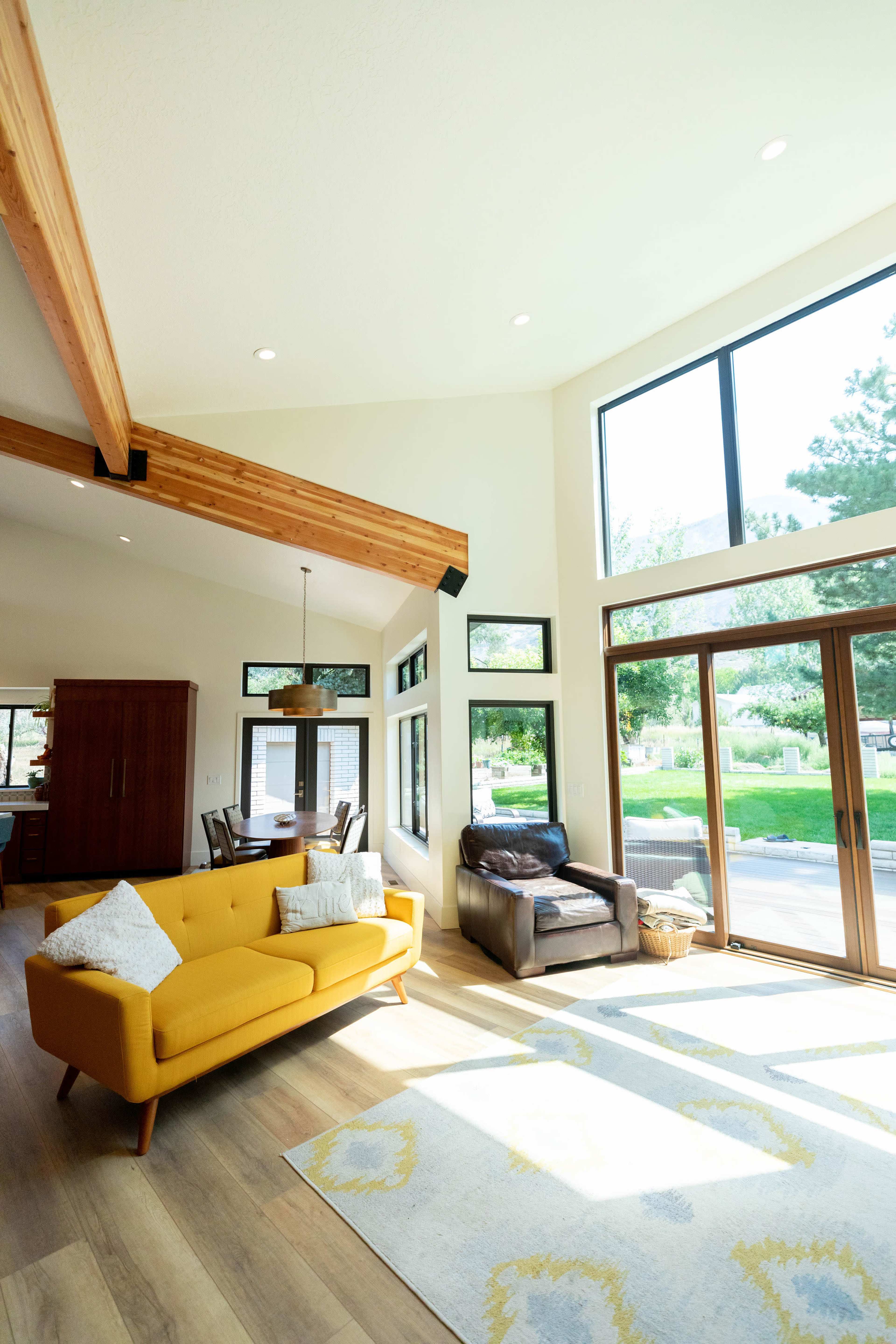 A bright living room features a yellow couch, a wooden dining table, large windows, and an open layout.