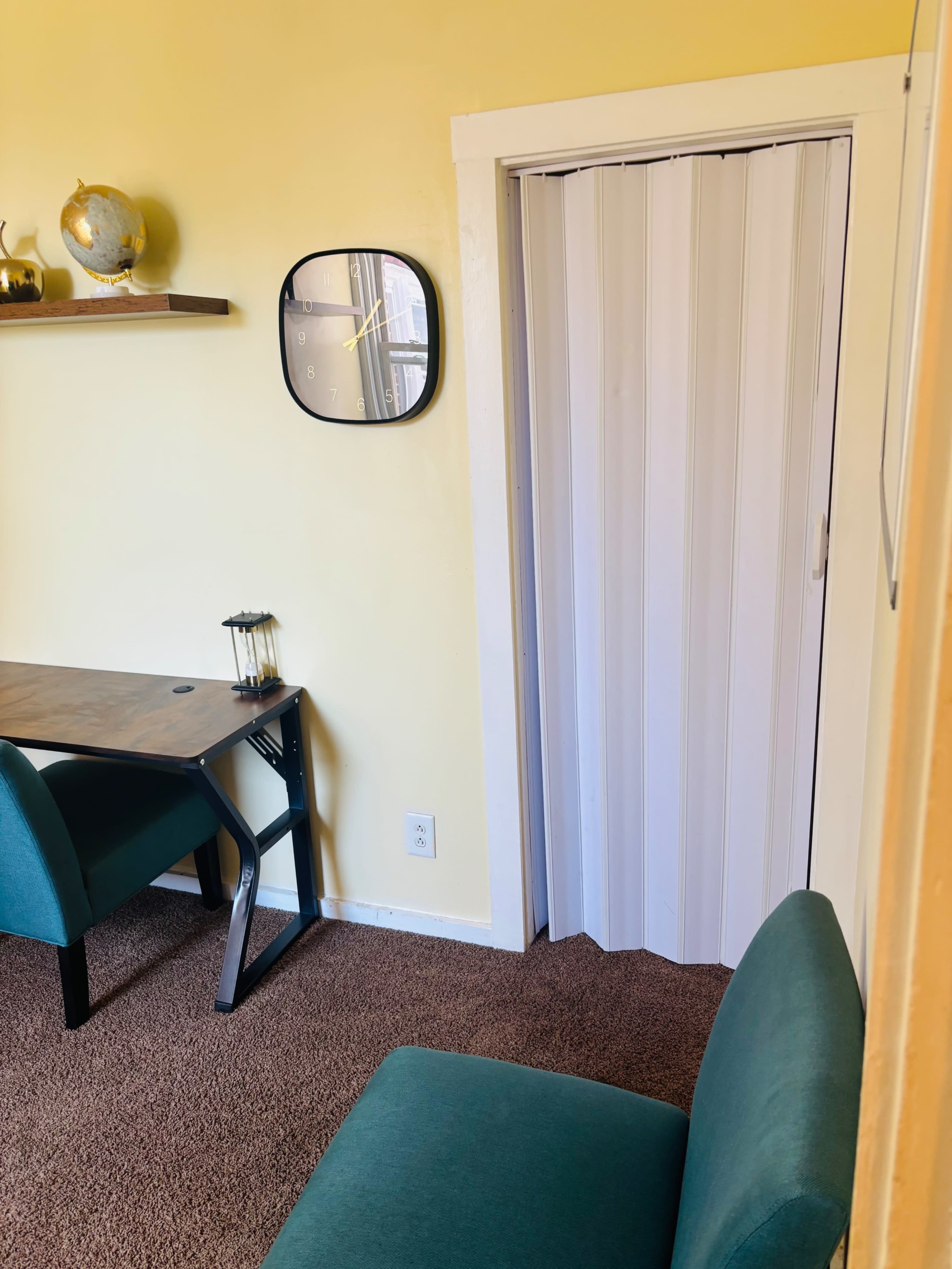 The image shows a small room with a yellow wall, a desk and two green chairs, and a closed folding door.