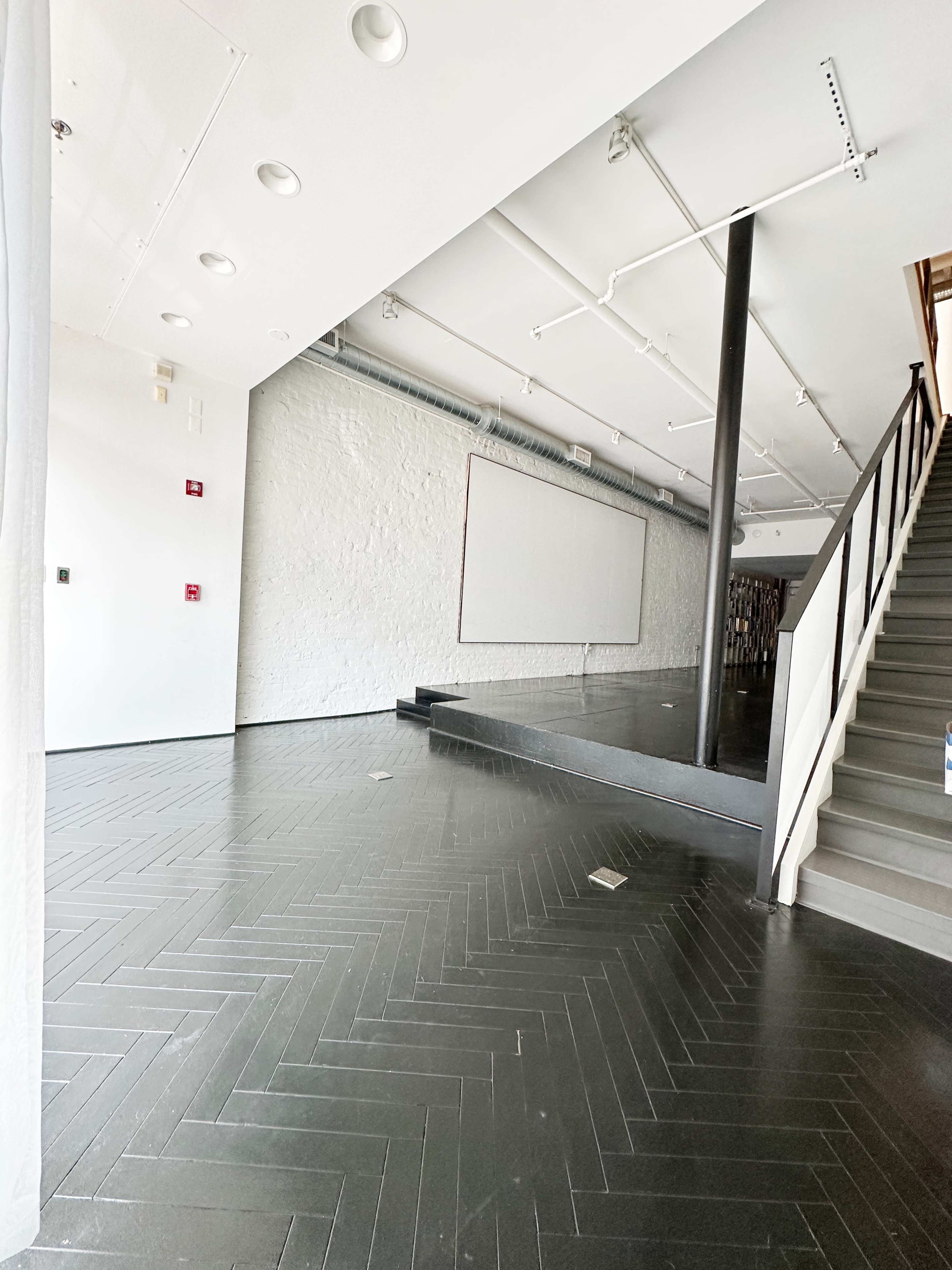 The image shows a spacious, empty interior with herringbone-patterned flooring, a staircase, and a blank wall at the far end.