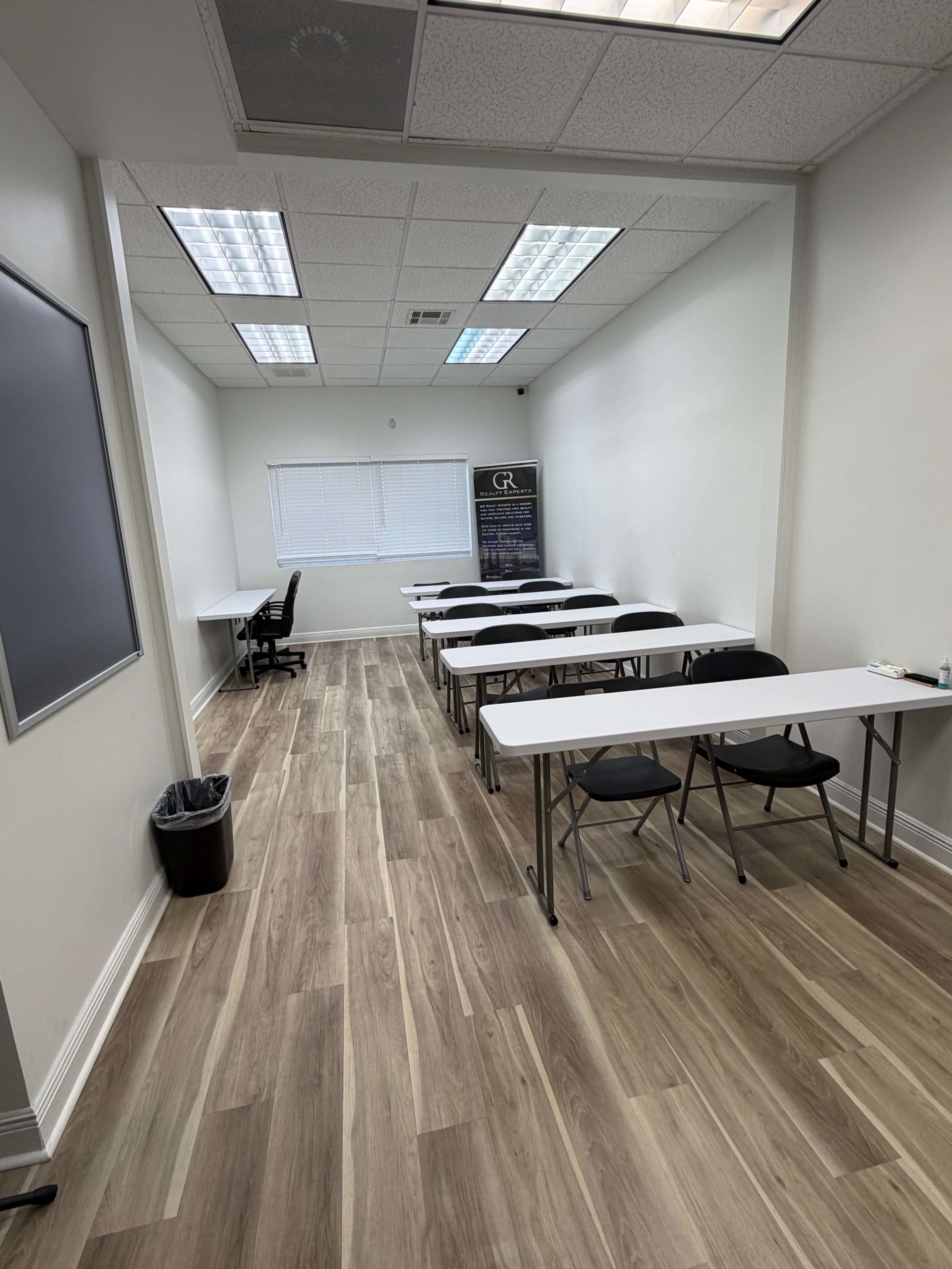 The image shows a sparsely furnished classroom with white tables and chairs arranged in two rows, wooden flooring, and a window allowing natural light.