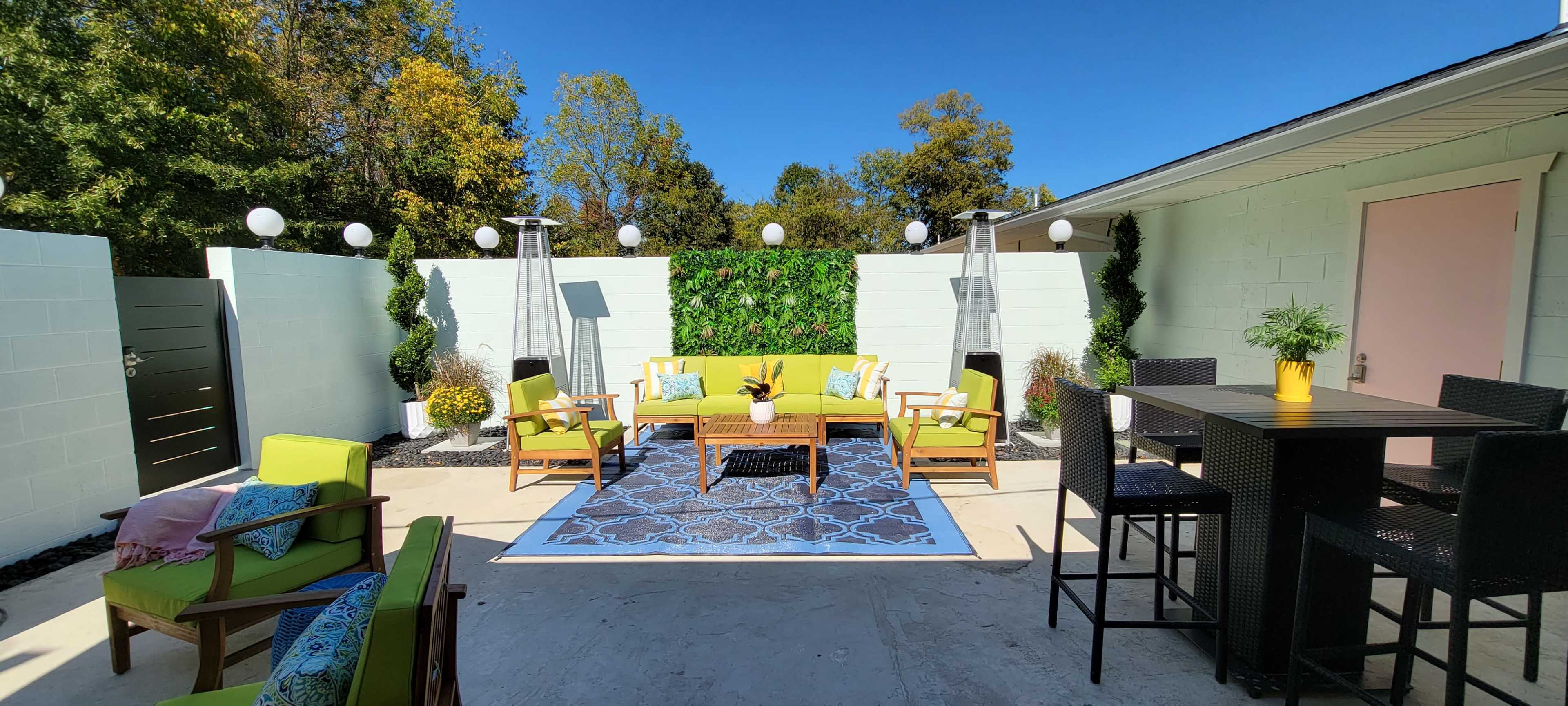 The outdoor space features a bright yellow sofa and chairs arranged around a patterned rug, flanked by decorative plants and lighting, with a patio dining set nearby.