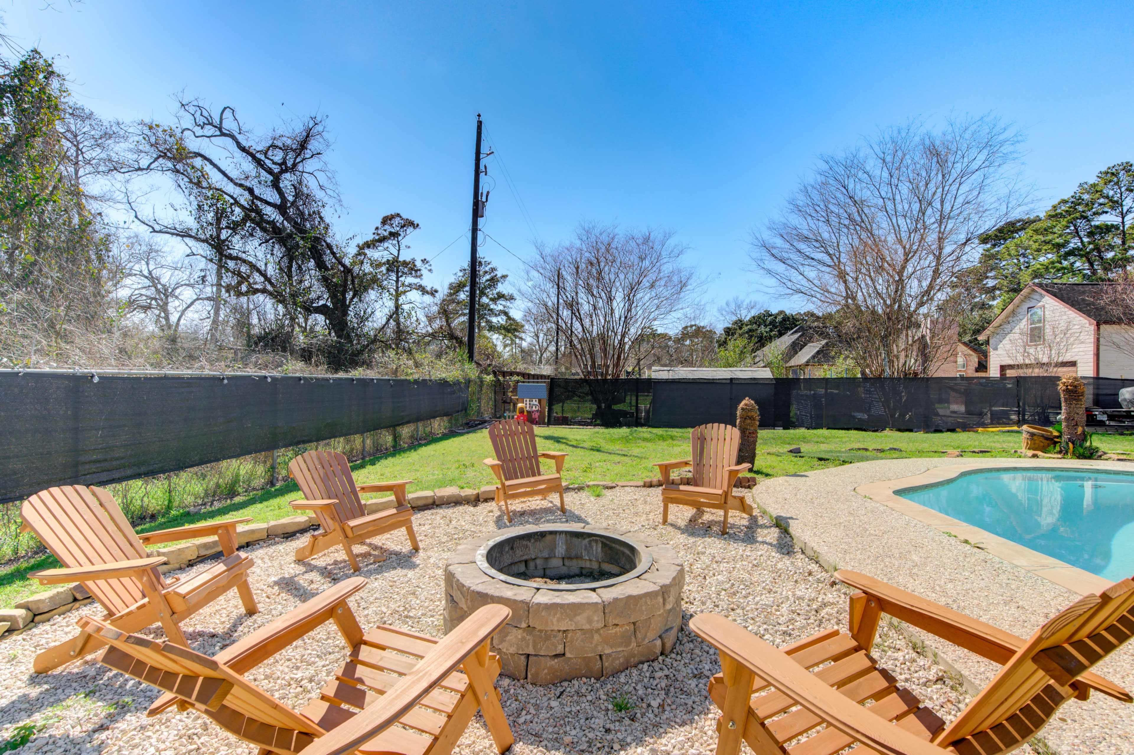 A circular stone fire pit surrounded by wooden adirondack chairs is situated next to a swimming pool in a grassy backyard.