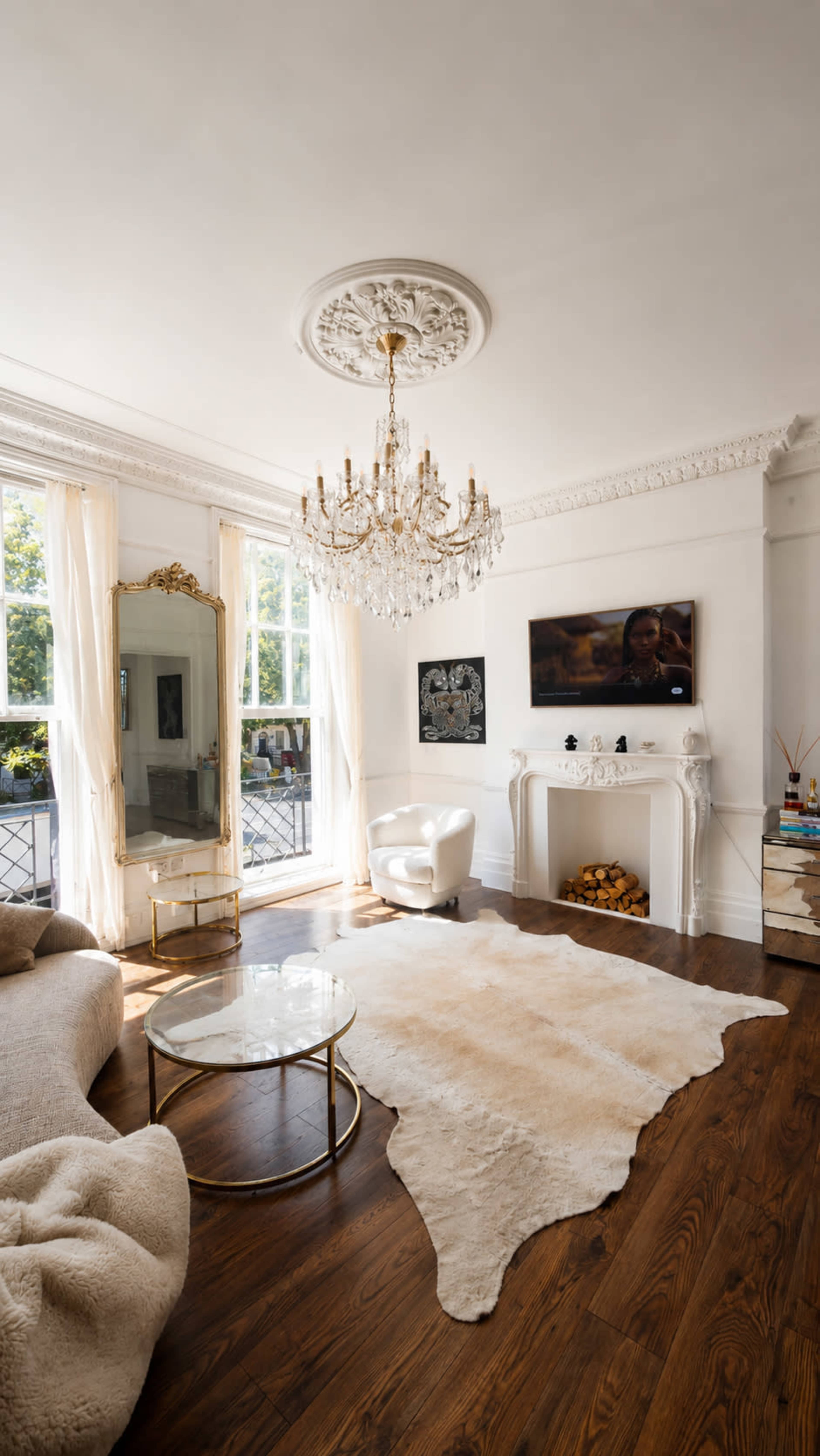 The living room features a chandelier, a large mirror, a modern white armchair, a circular coffee table, and a decorative fireplace, all arranged on a wooden floor with a cowhide rug.