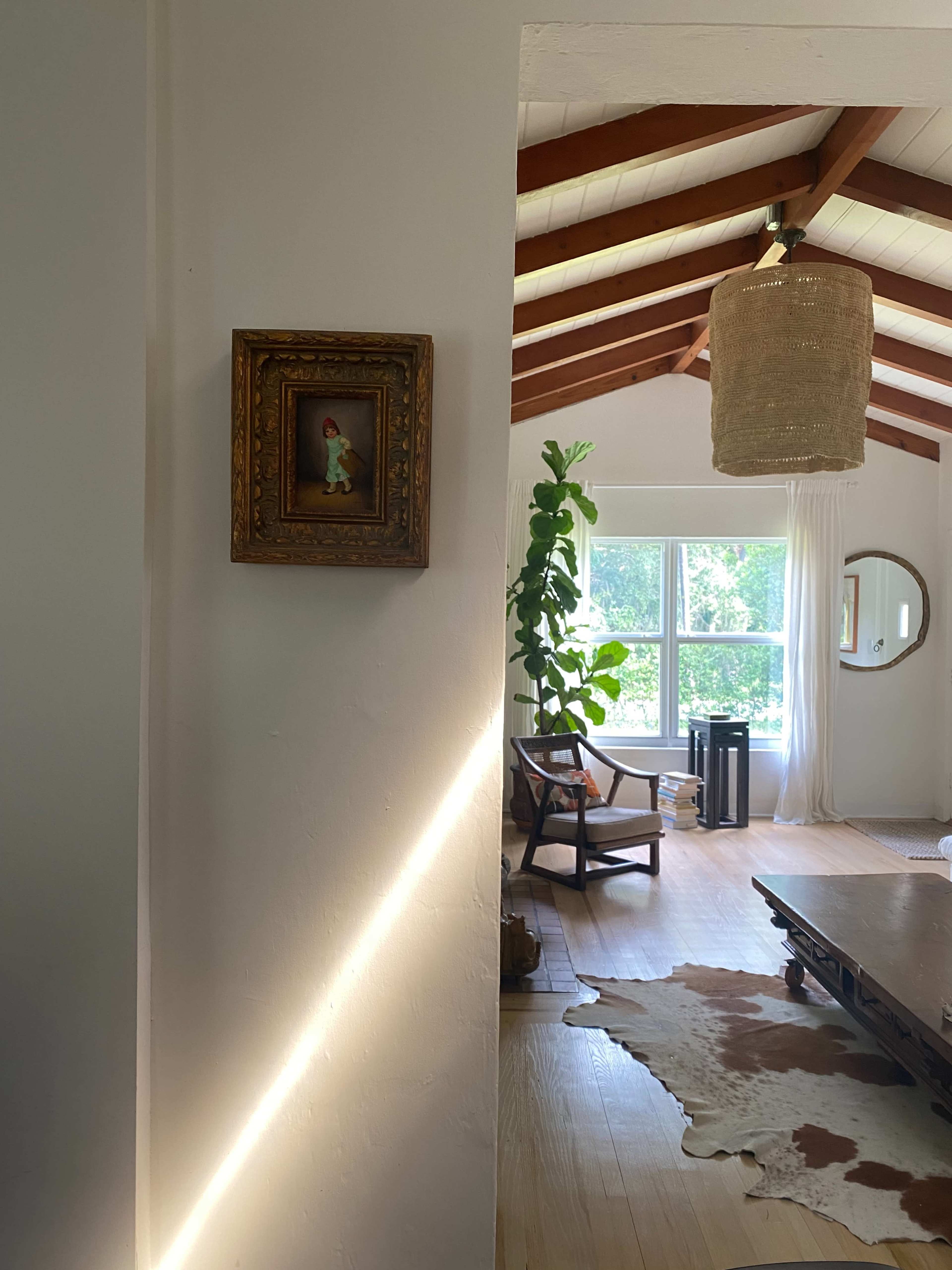 A framed artwork hangs on a white wall beside a sunlit living room featuring a large window, a green plant, and a wooden table.