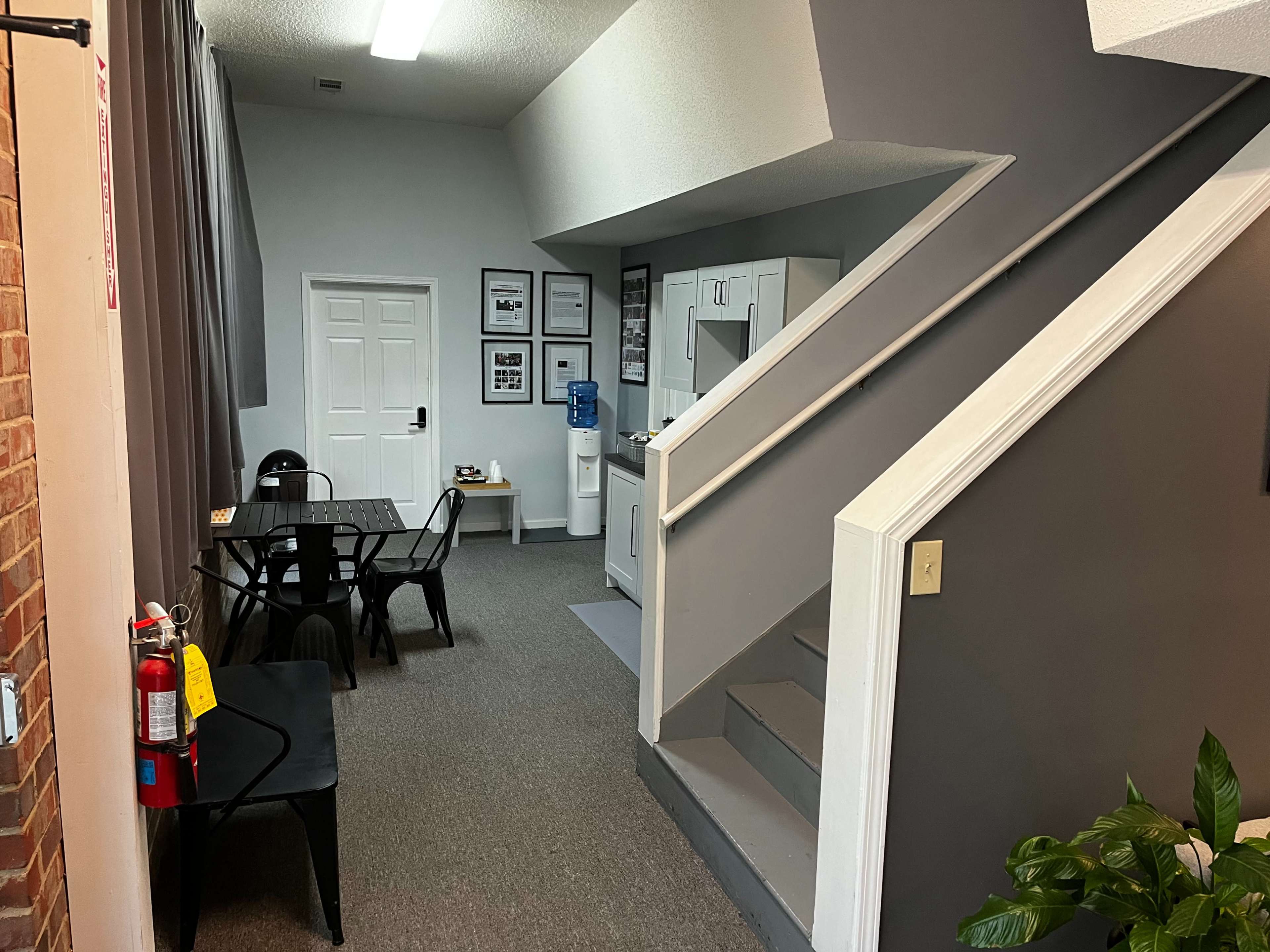 The image shows a small interior space featuring a staircase, a kitchenette area, a dining table with chairs, and gray walls.