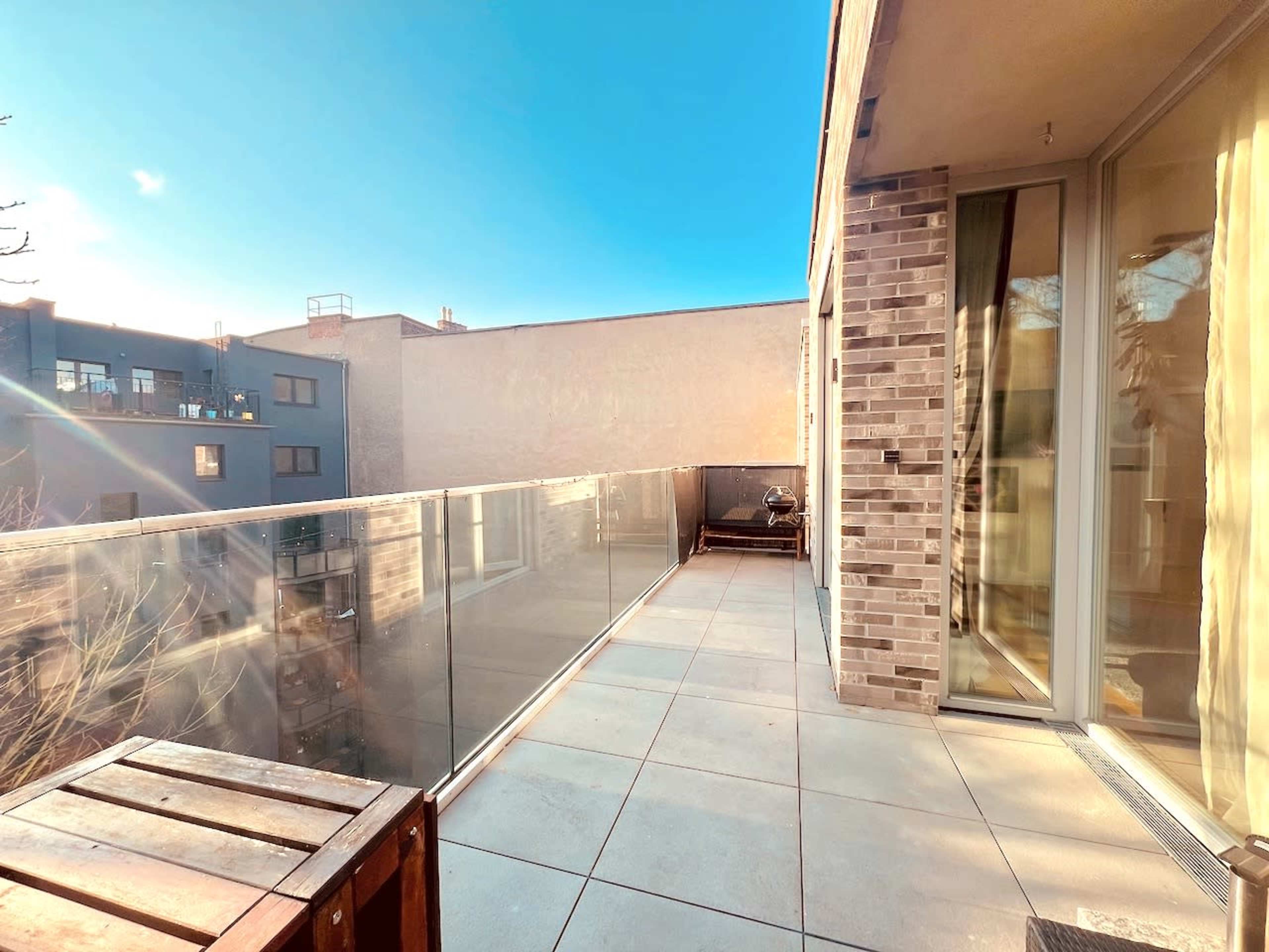 The image shows a spacious balcony with a glass railing, tiled floor, and a view of neighboring buildings under a clear blue sky.