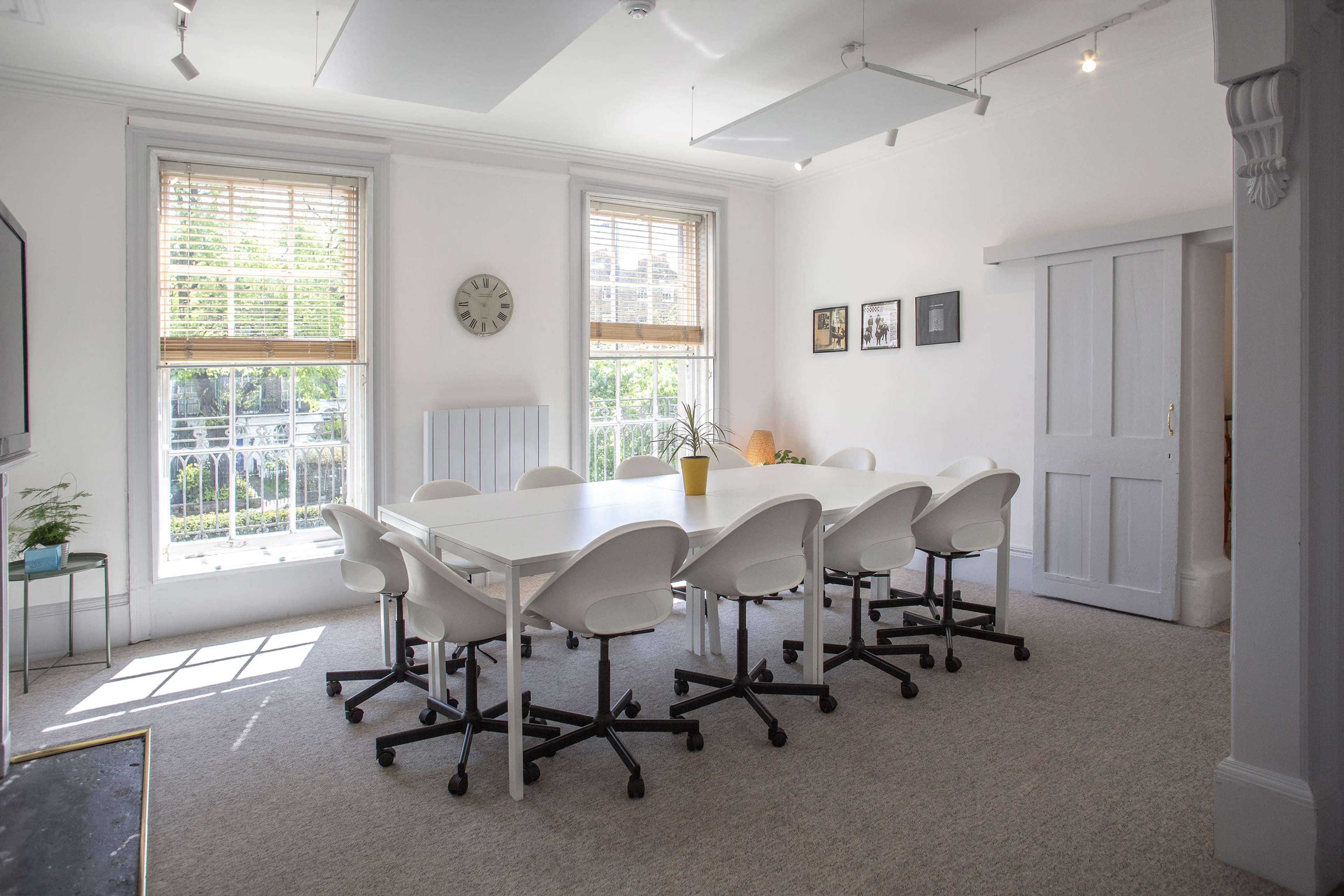 A brightly lit meeting room features a large white table surrounded by eight white chairs, with windows offering a view of greenery outside.