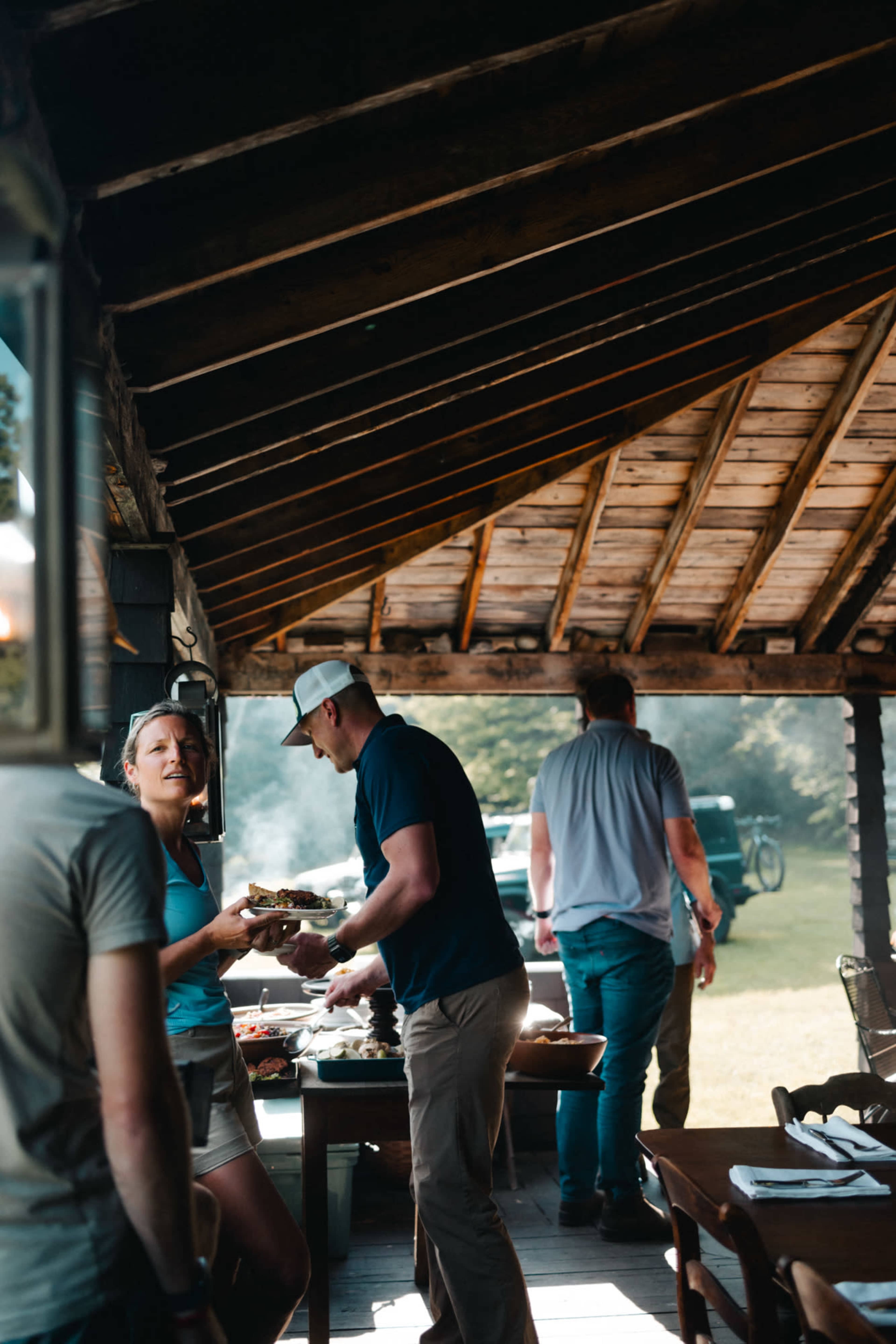 A group of people gather inside a rustic cabin, sharing food while another individual steps outside.