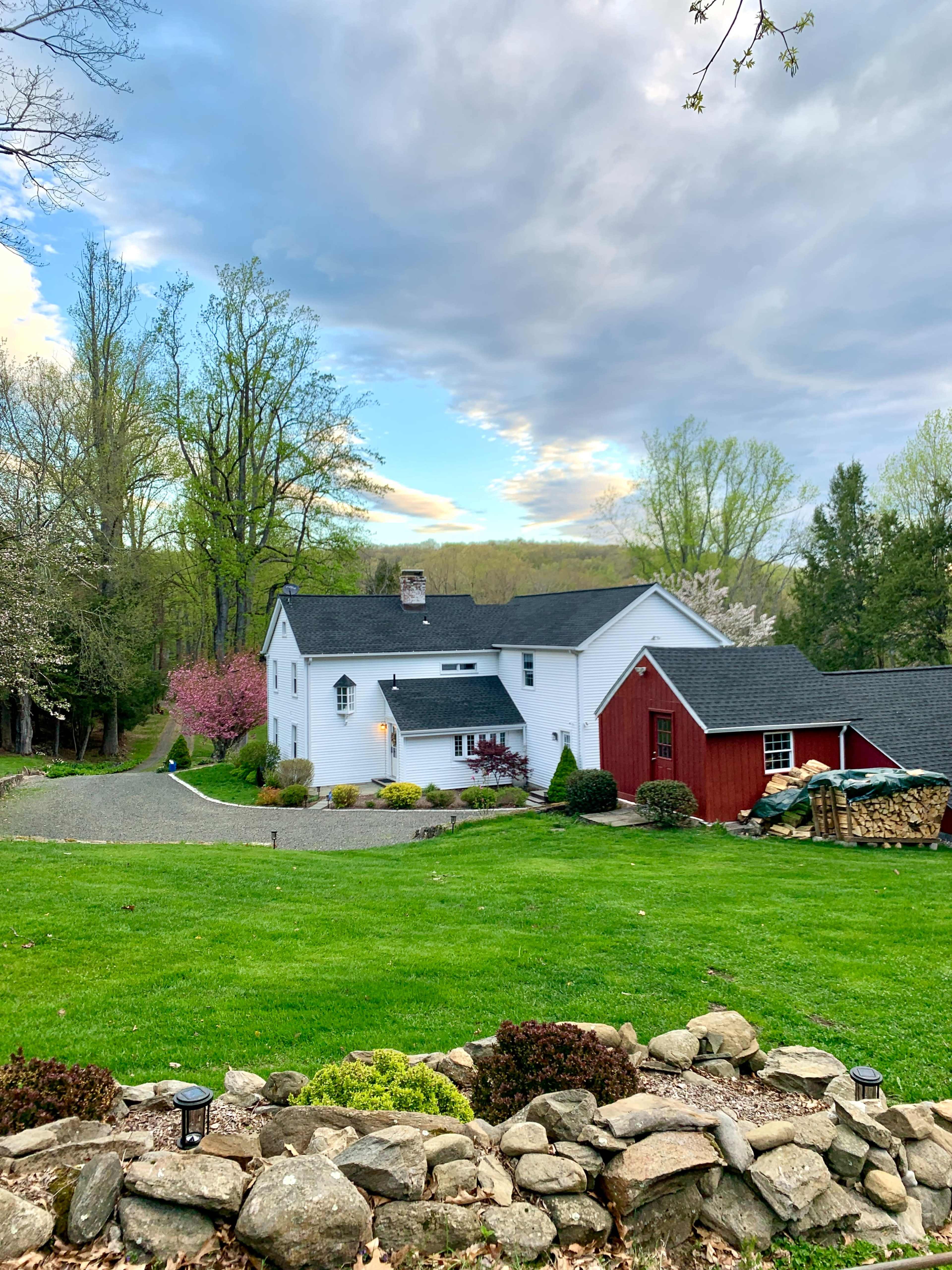 A white house with a red barn-style garage is surrounded by green grass and trees, set against a cloudy sky.