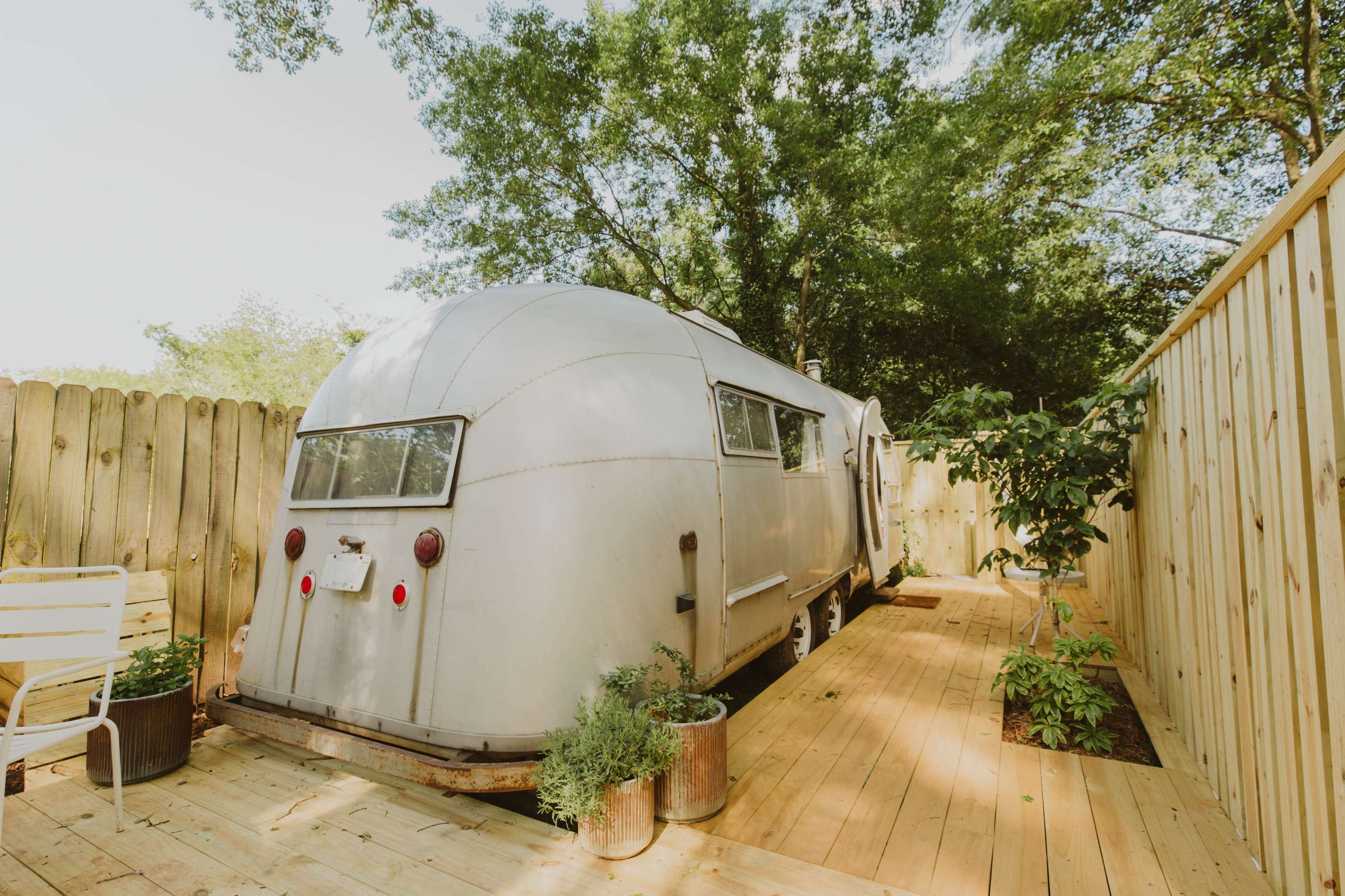 A vintage camper is parked on a wooden deck surrounded by a wooden fence and greenery.