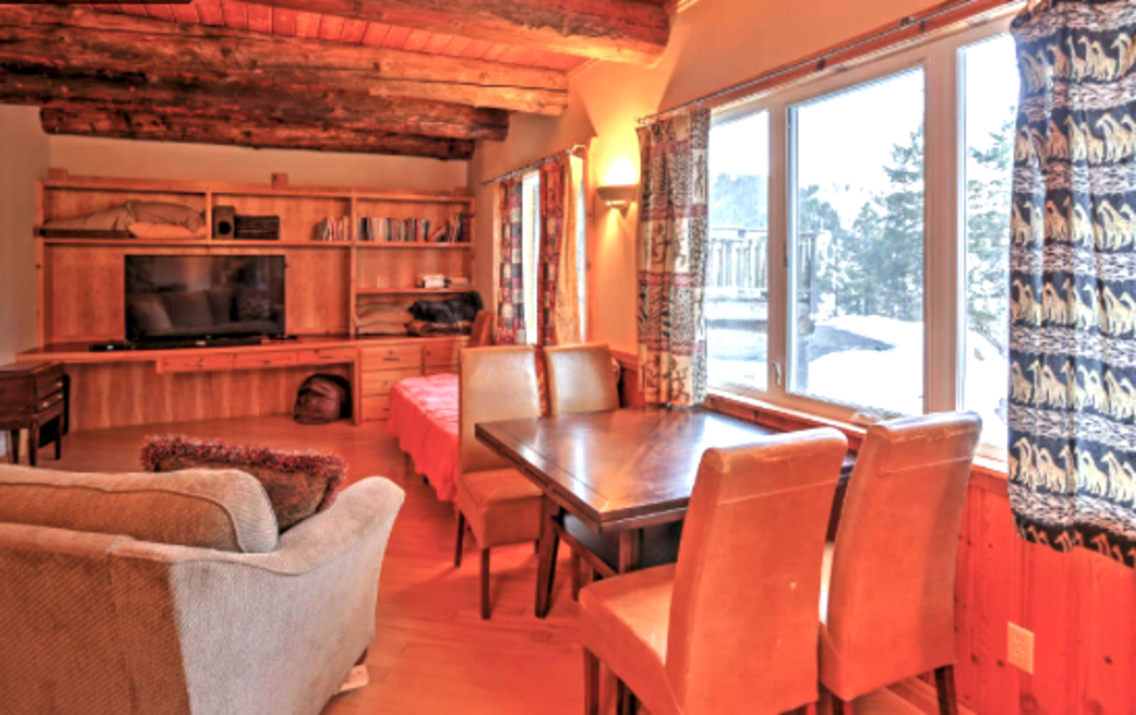 A cozy living room with wooden beams, a television on a shelf, a dining table with four chairs, and large windows allowing natural light.