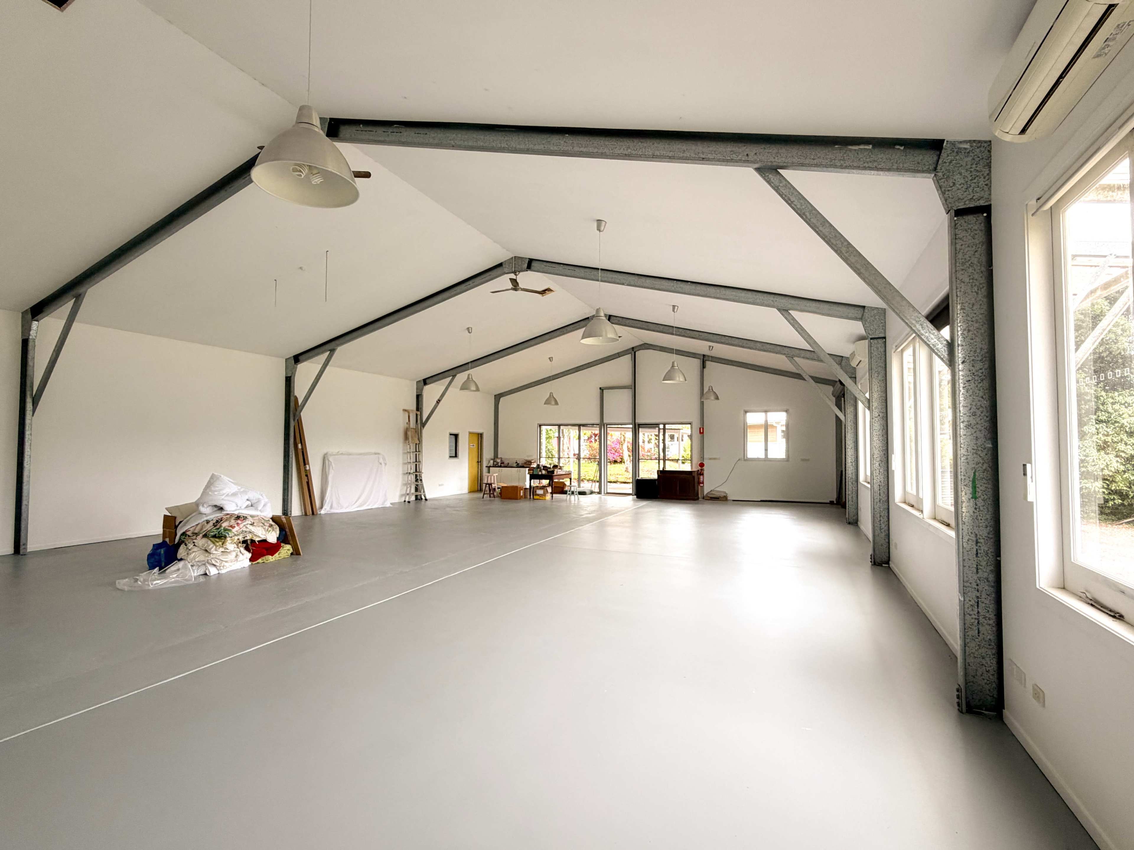The image shows a spacious indoor area with high ceilings, metal beams, minimal furniture, and large windows displaying greenery outside.