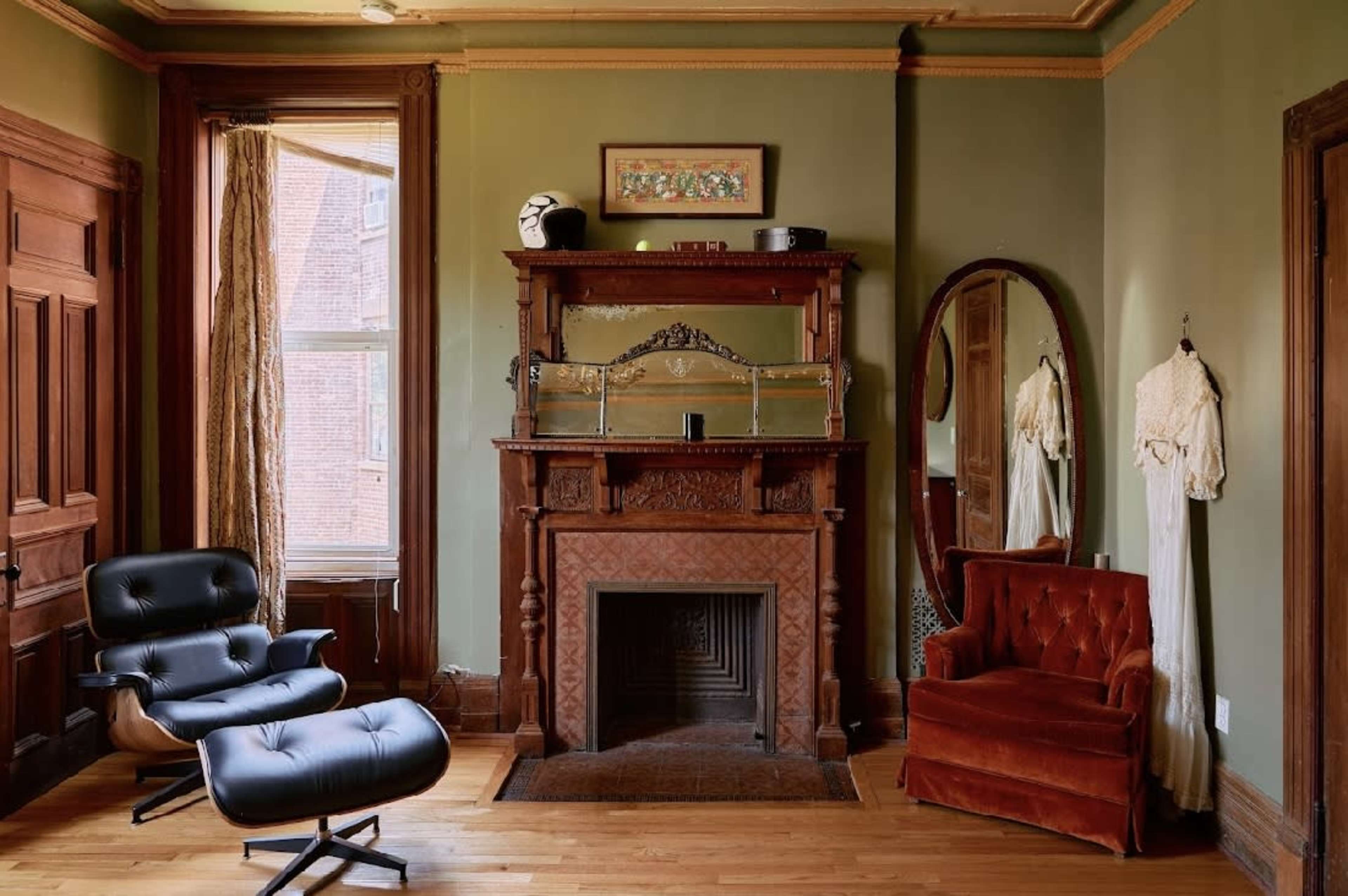 The image shows a vintage-style room with a wooden fireplace, a dark upholstered chair, a red velvet sofa, and a large mirror on the wall.