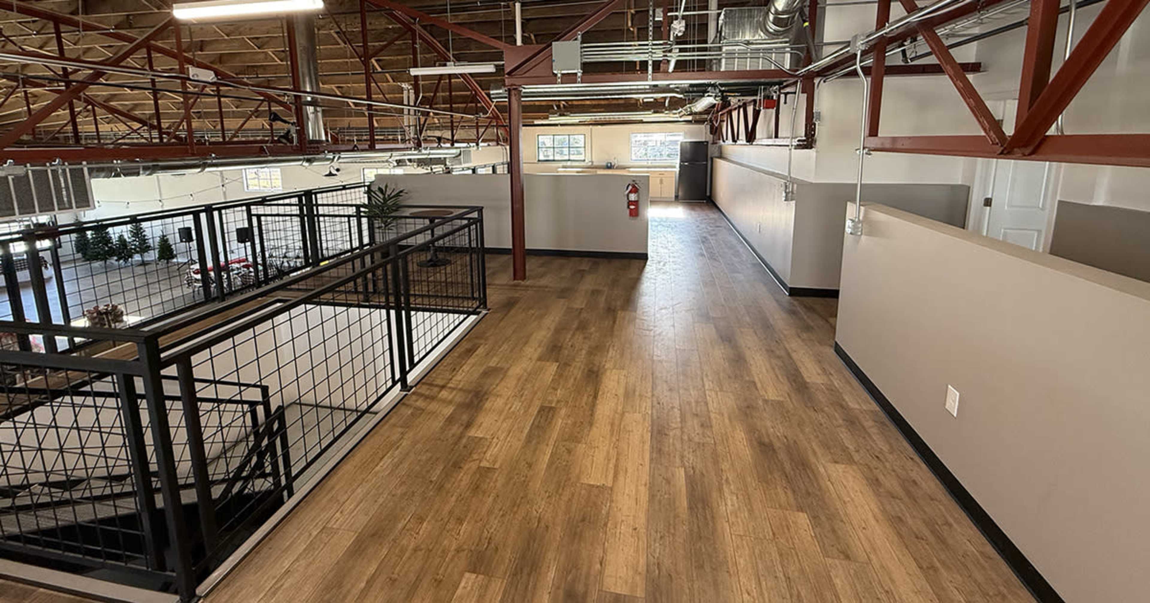 The image shows a spacious interior hallway with wood flooring, metal railings, and exposed structural beams, leading to an open area with natural light.