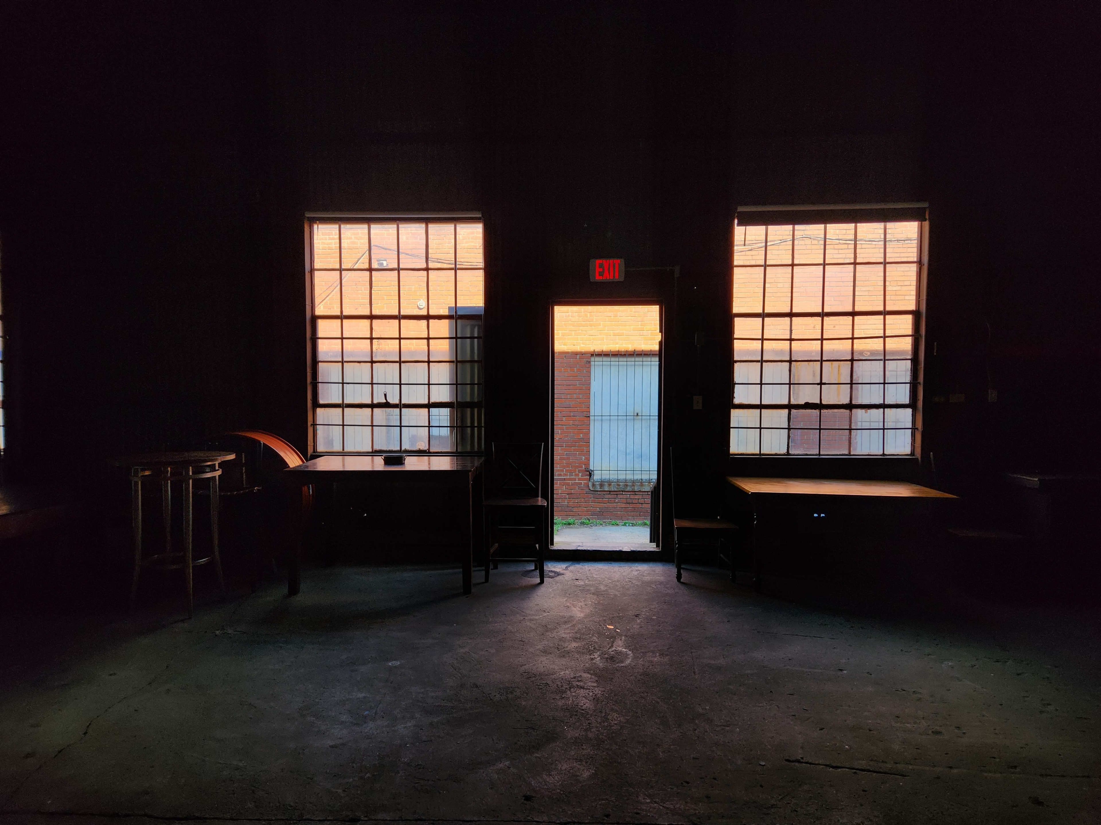 A dimly lit room with two large windows and an open door leading outside to a brick wall and a grassy area.