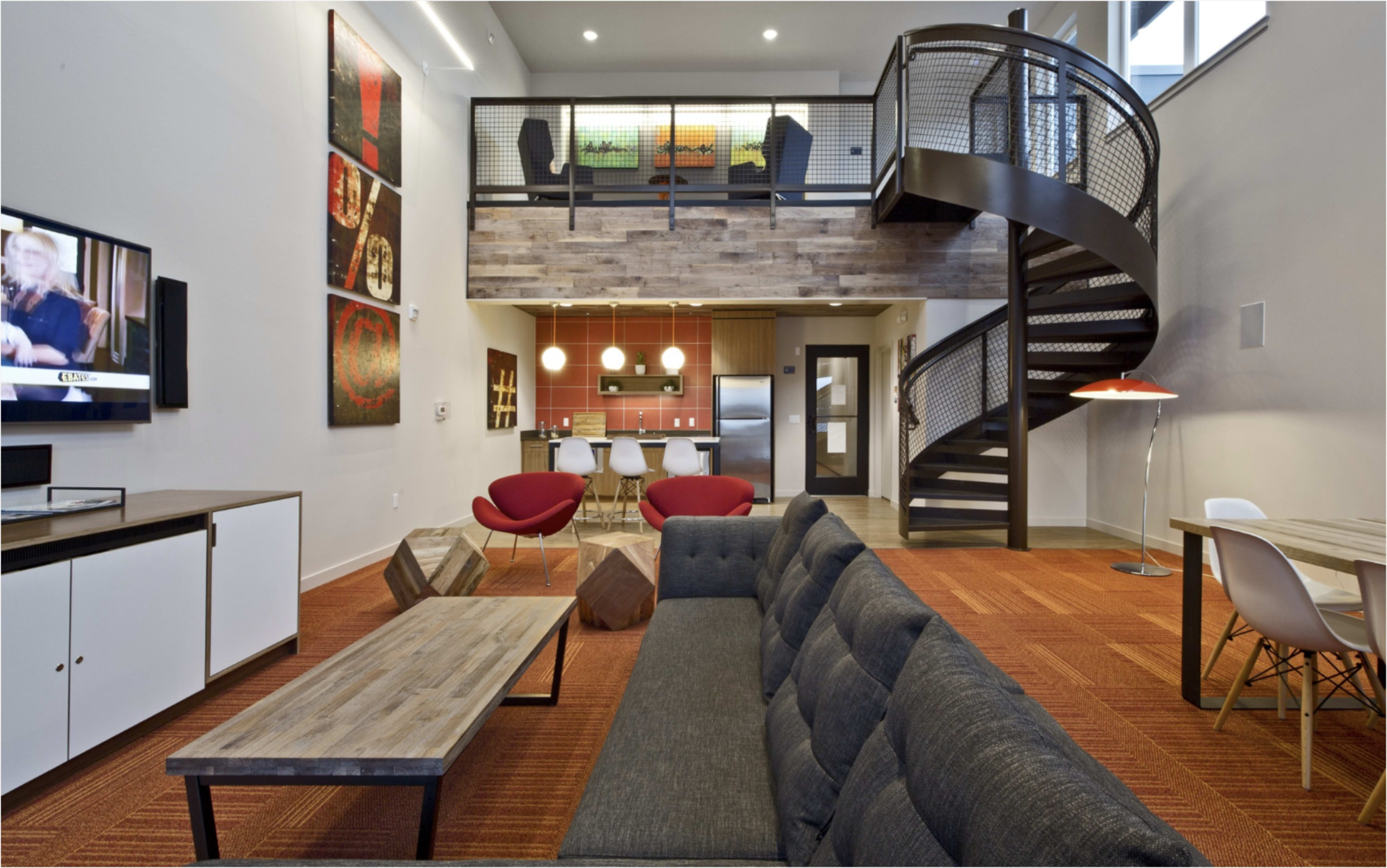 The image shows a modern, open-concept living space featuring a staircase, a kitchen area with bar stools, and a cozy seating arrangement with a dark sofa and two red chairs.