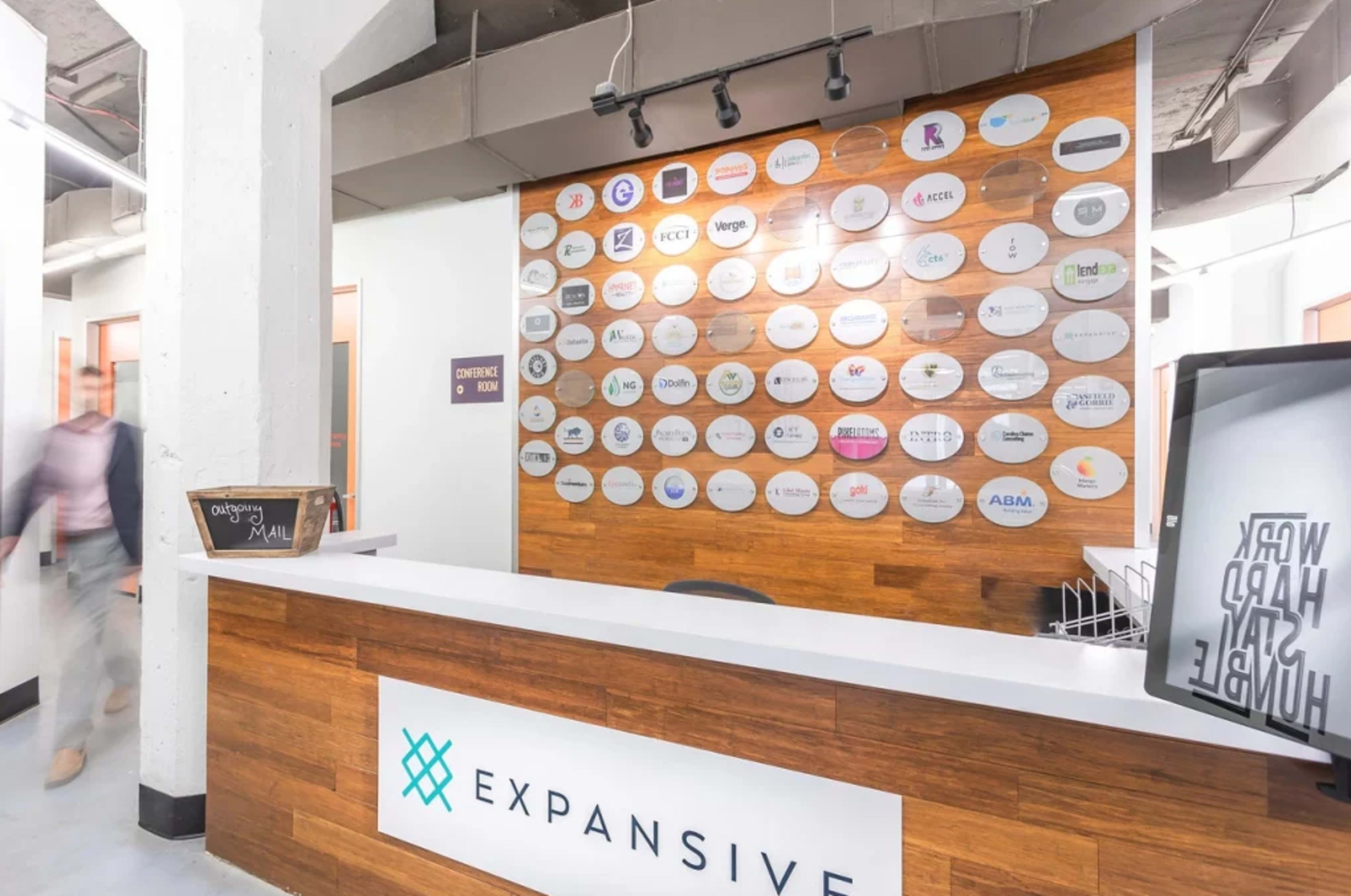 The image shows a modern reception area with a wooden wall displaying multiple company logos and a front desk labeled "EXPANSIVE."