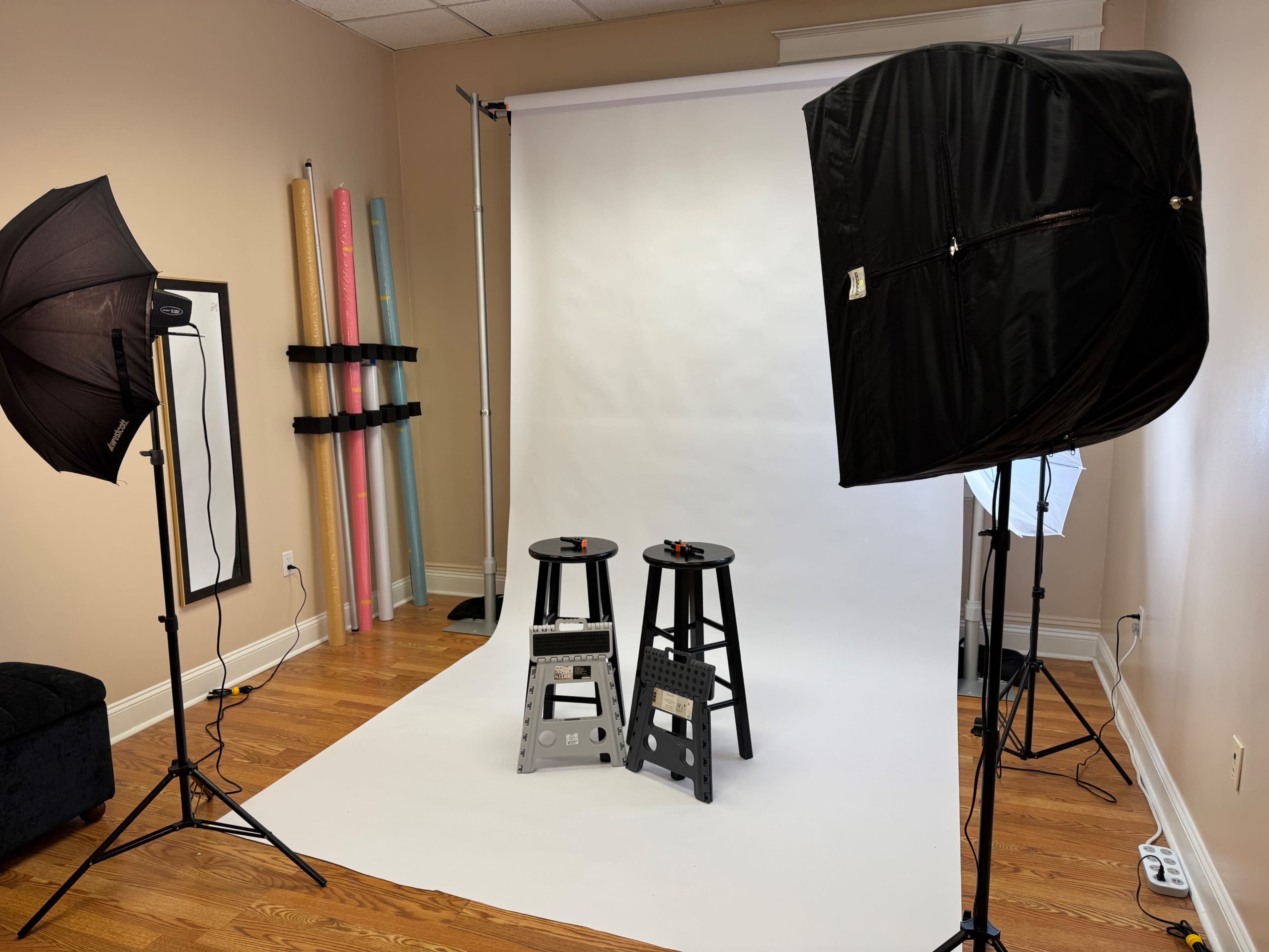The image shows a photography studio setup with two stools positioned on a white backdrop, surrounded by softbox lights and colorful rolls of paper along the wall.