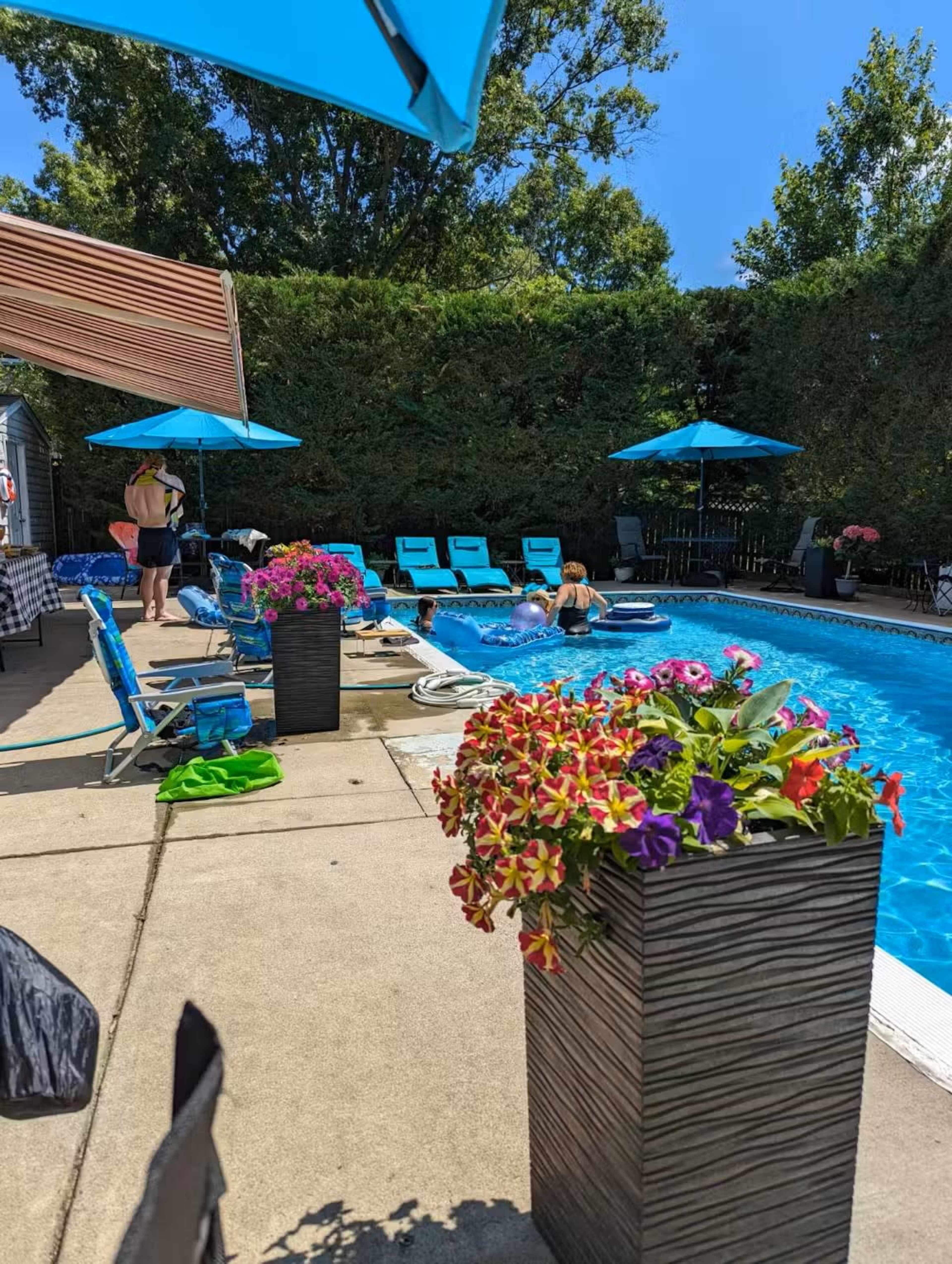 The scene shows a swimming pool area with sun loungers, vibrant flower planters, and several umbrellas providing shade.