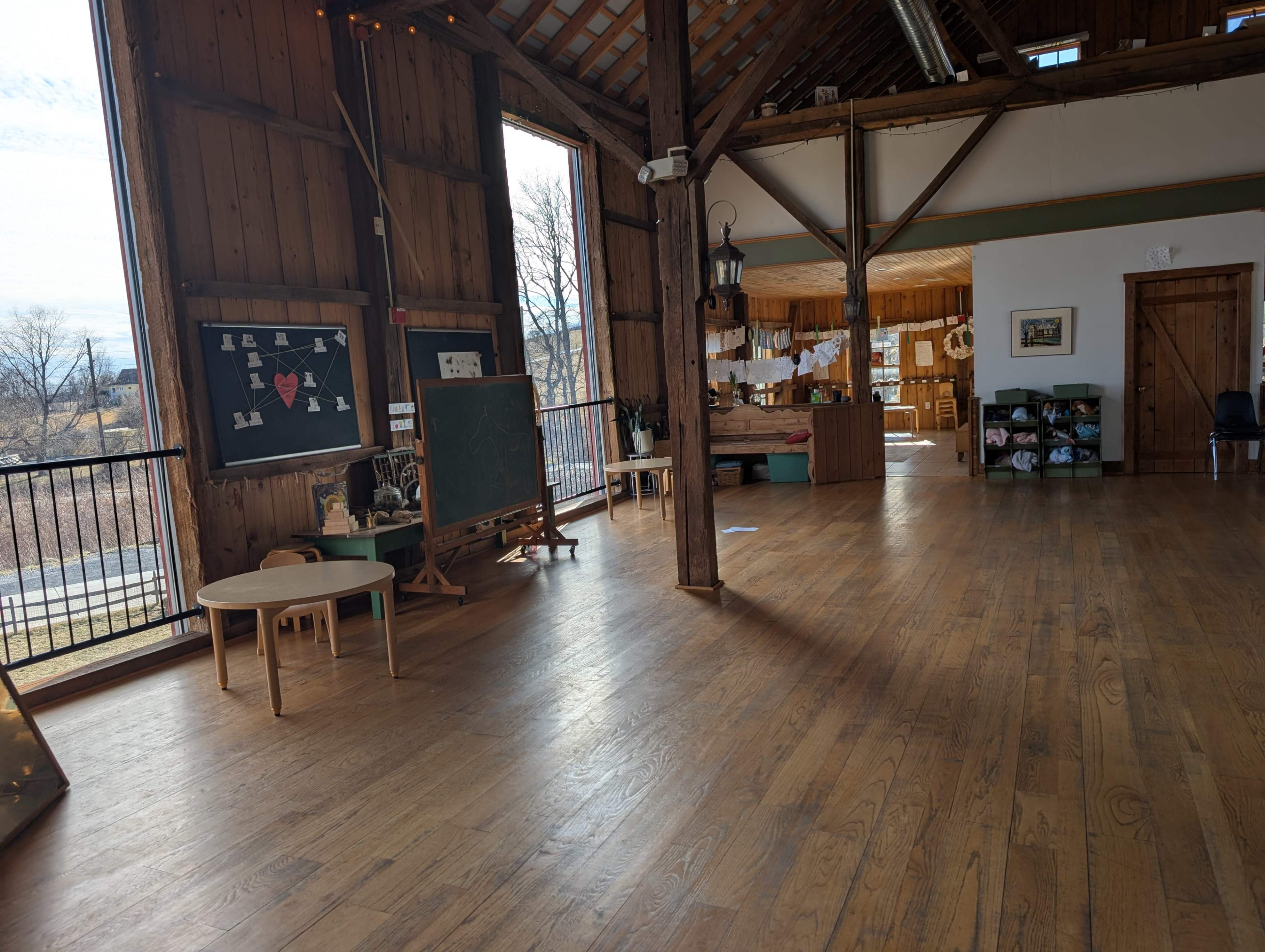 The image shows a spacious wooden interior featuring a large open area with tables, a chalkboard, and a kitchenette visible in the background.