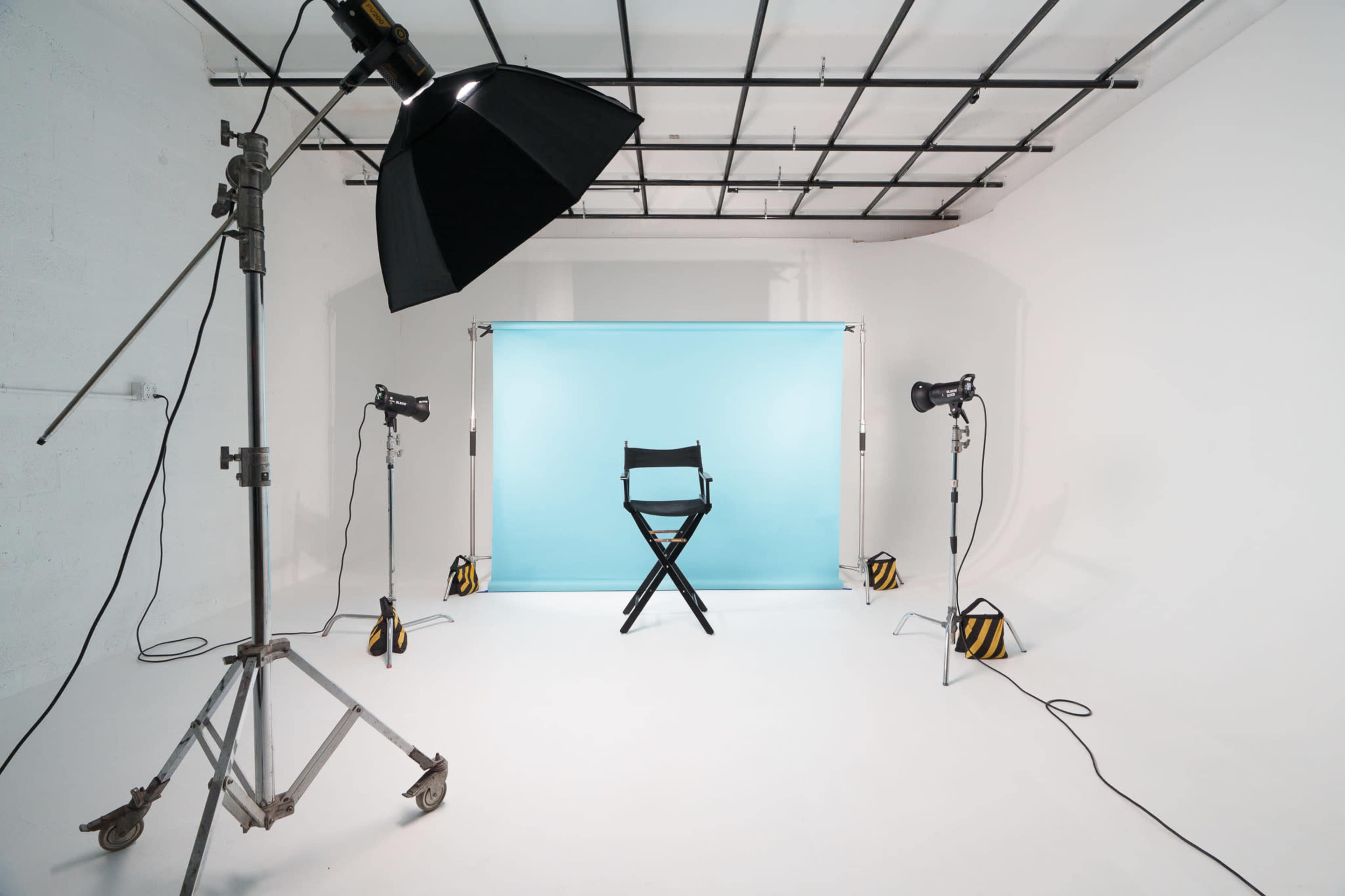 A photography studio features a black director's chair in front of a light blue backdrop, surrounded by studio lights and equipment.