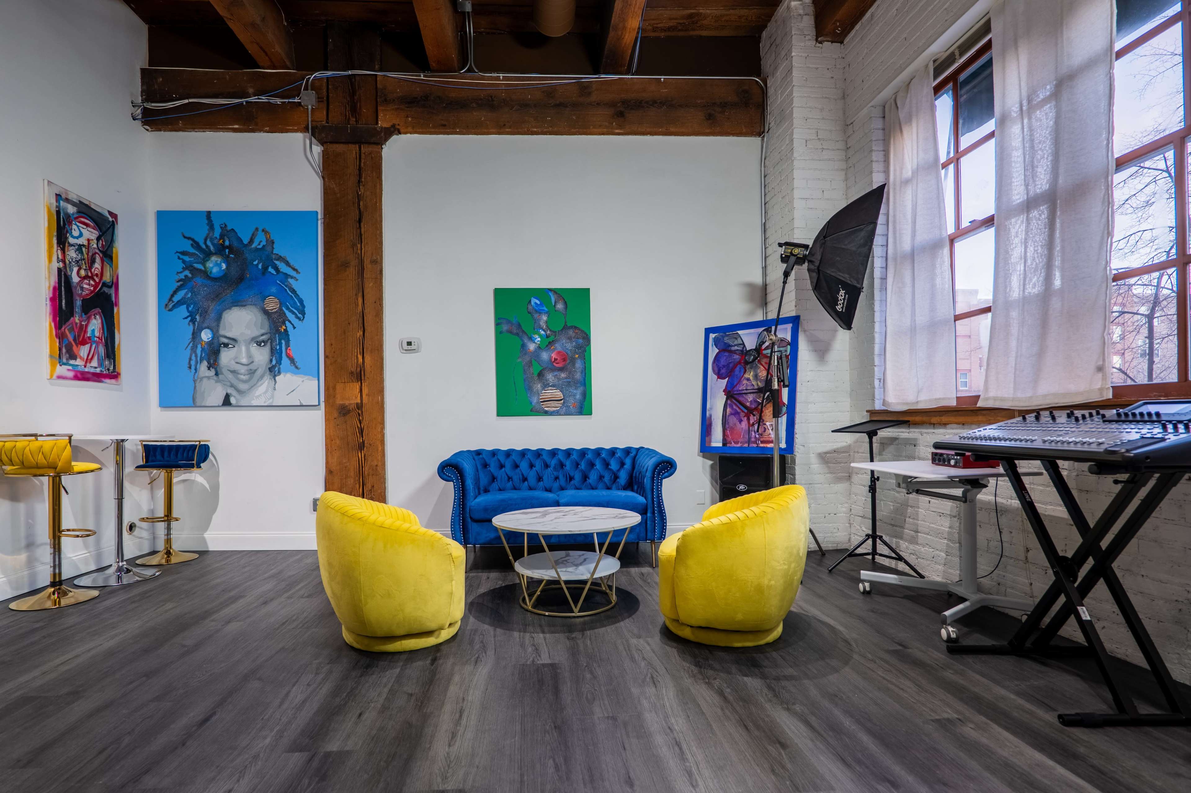 The scene features a modern, open living space with a blue tufted sofa, two yellow accent chairs, a round coffee table, colorful artwork on the walls, and a music production setup.