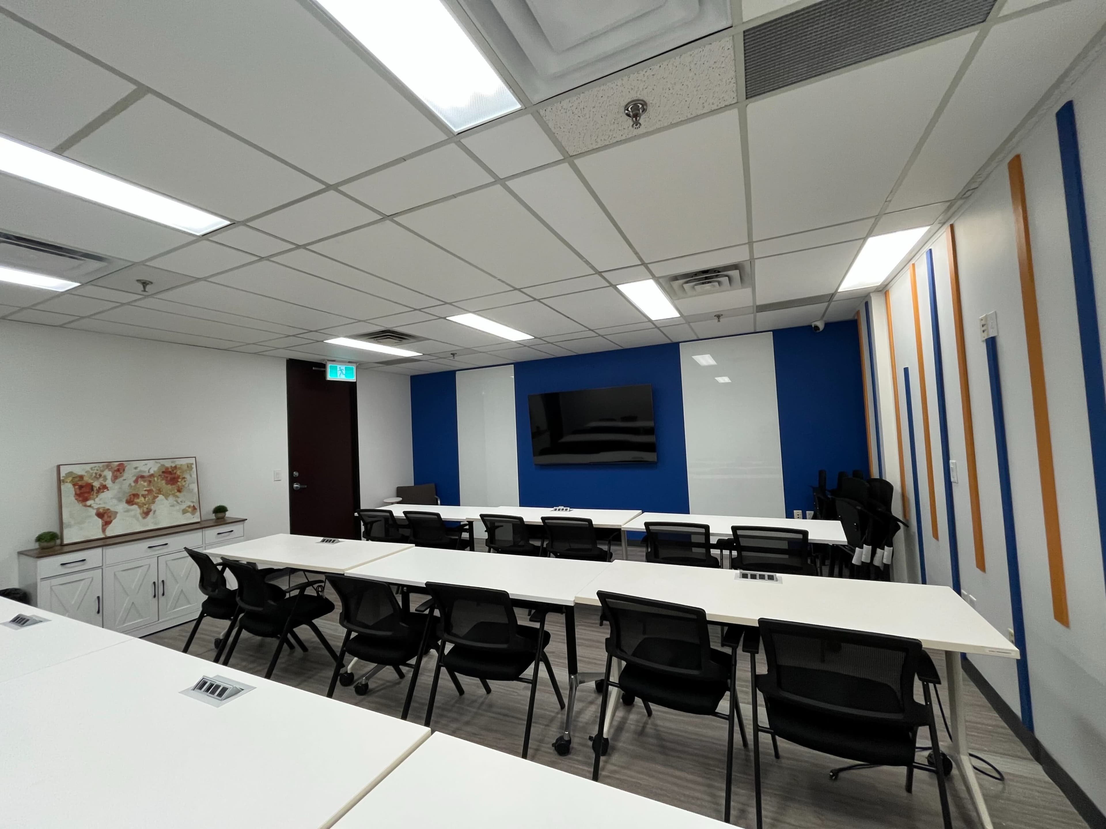 The image shows a modern conference room with multiple tables arranged in a U-shape, featuring a large screen on the wall, and equipped with chairs and a world map decoration.