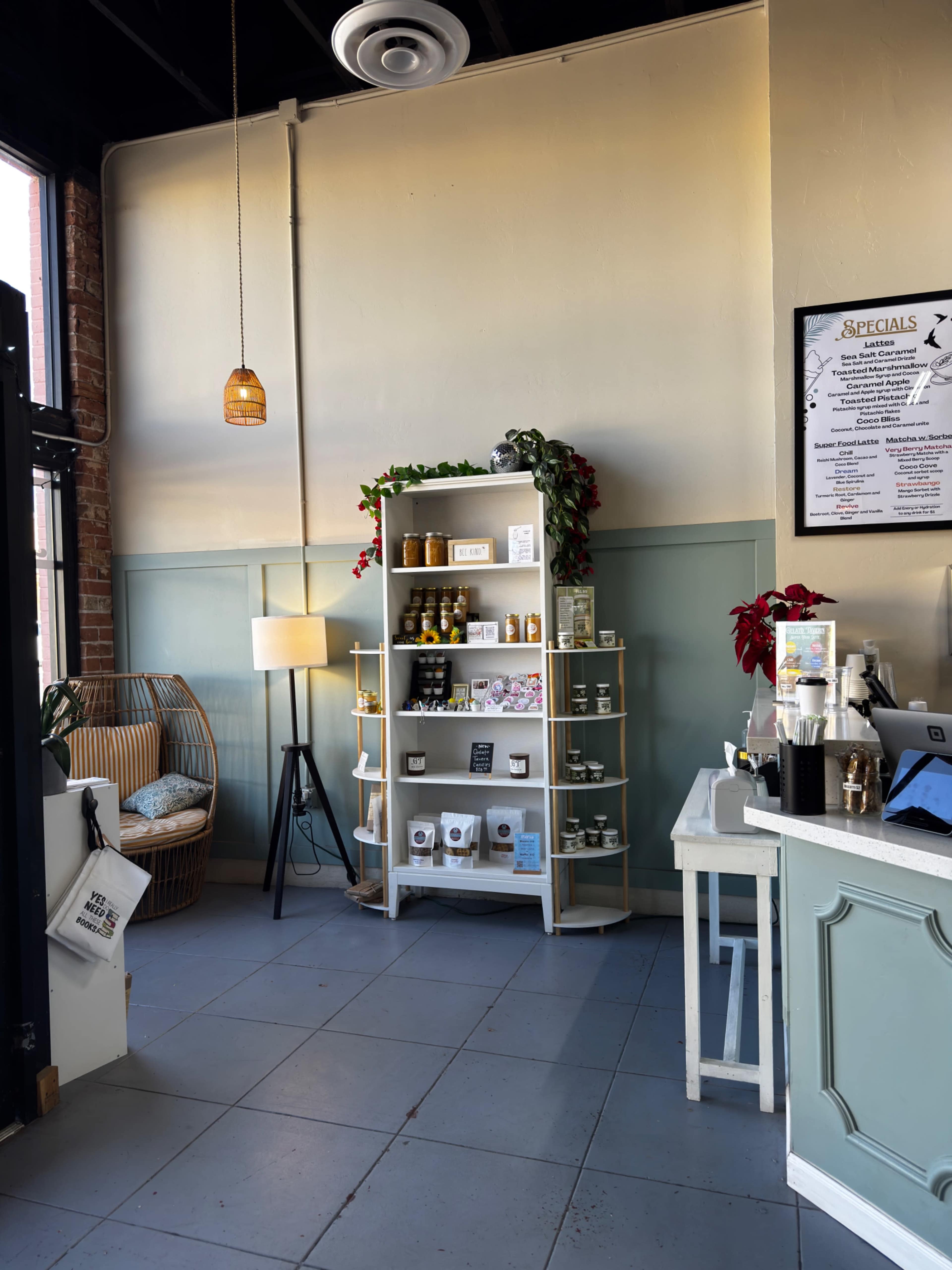 The image shows a cozy corner of a café featuring a shelf with various products, a lamp, and a comfortable chair.