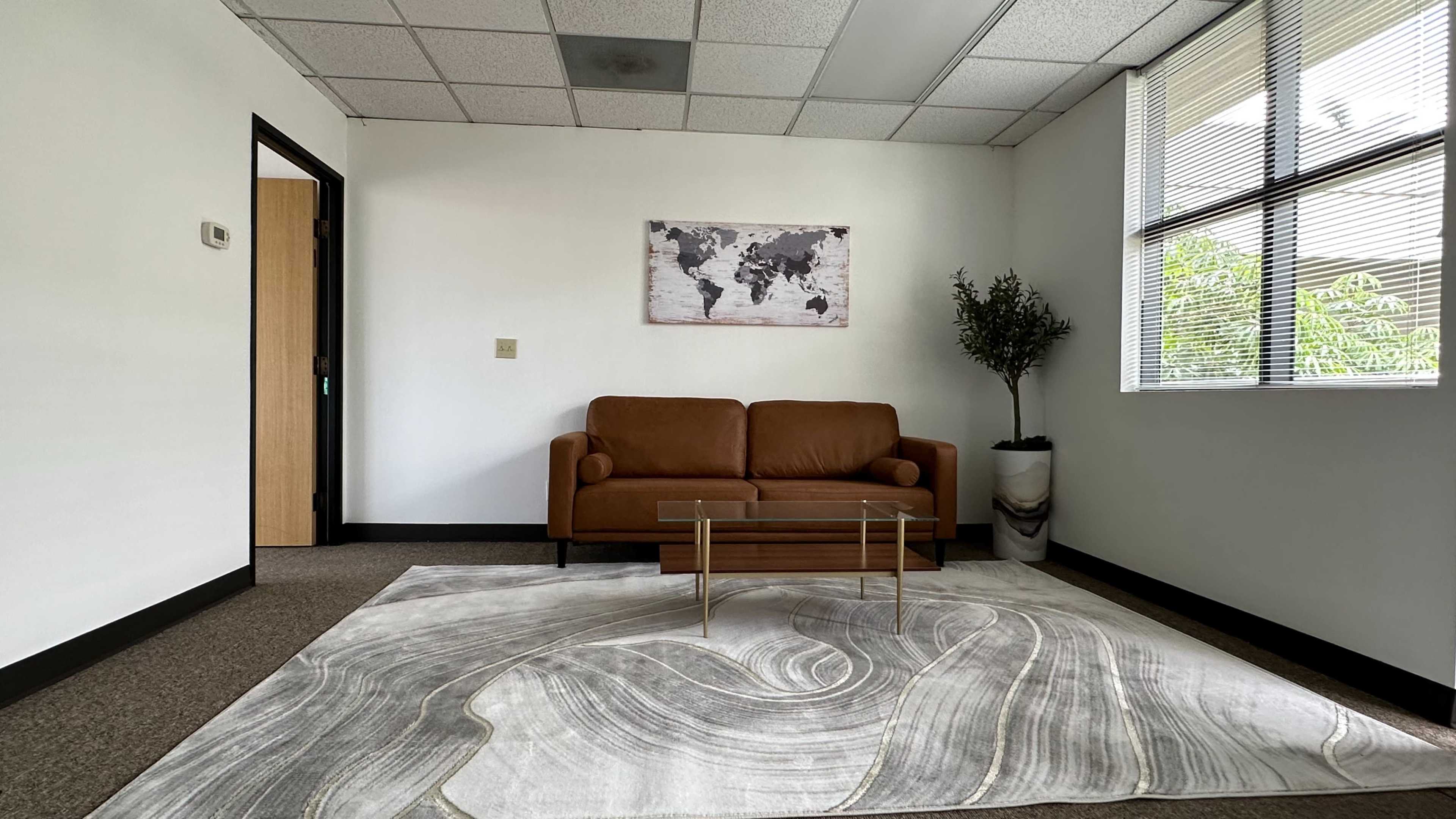 A modern office room features a brown sofa, a glass coffee table, and a world map on the wall, with a potted plant near a window.