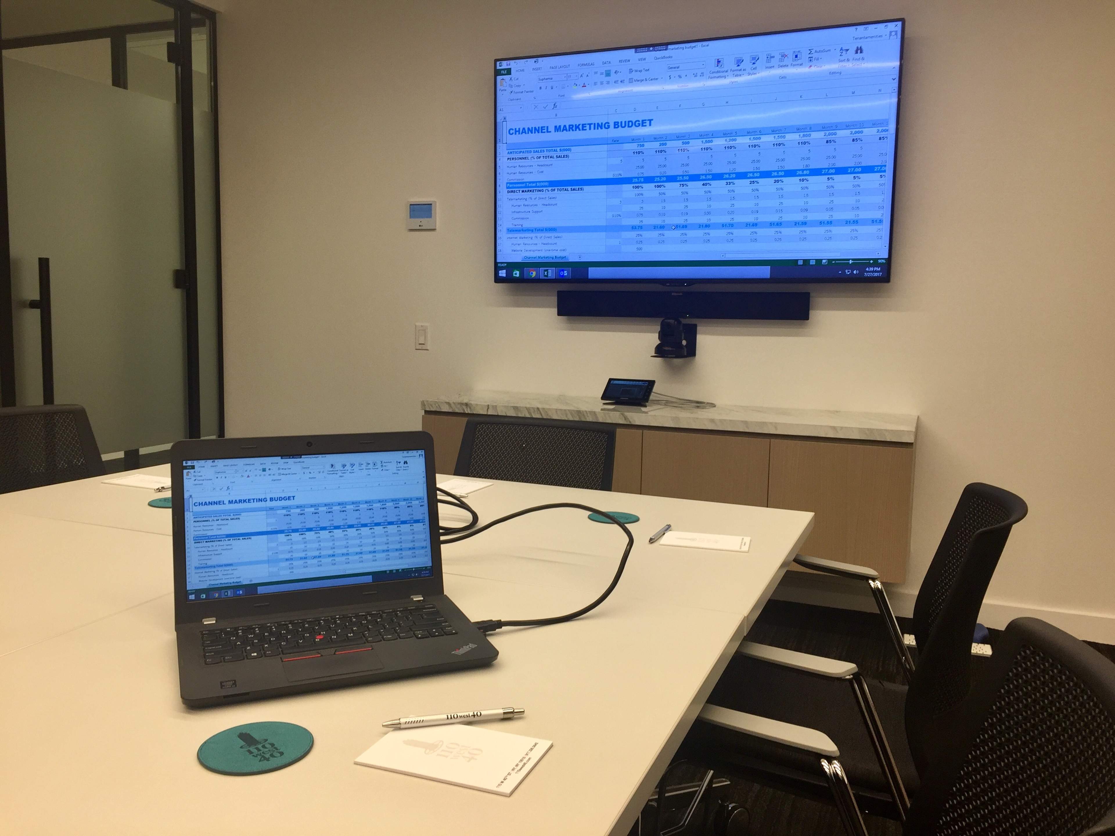 A laptop is placed on a conference table with an Excel spreadsheet on a large screen in the background, displaying a channel marketing budget.
