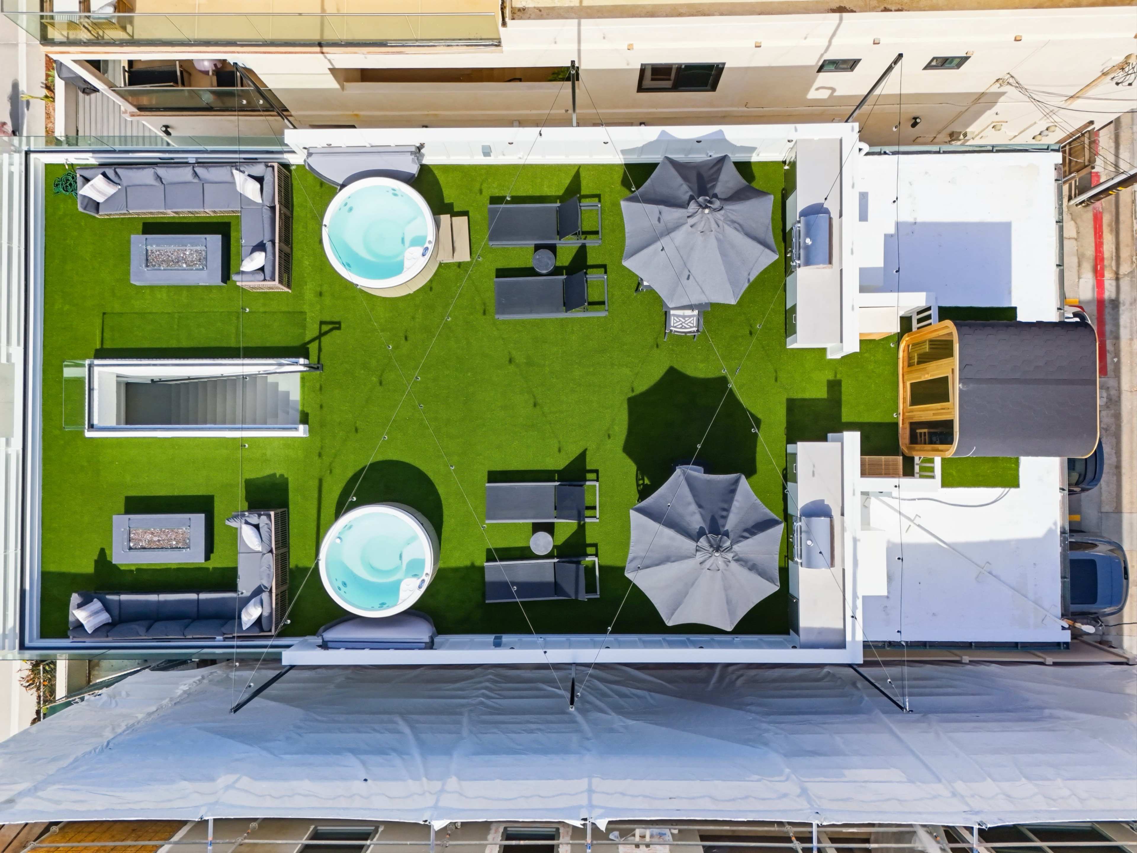 The image shows a rooftop terrace featuring two hot tubs, several seating areas with gray furniture, and multiple shaded umbrellas on a green artificial grass surface.