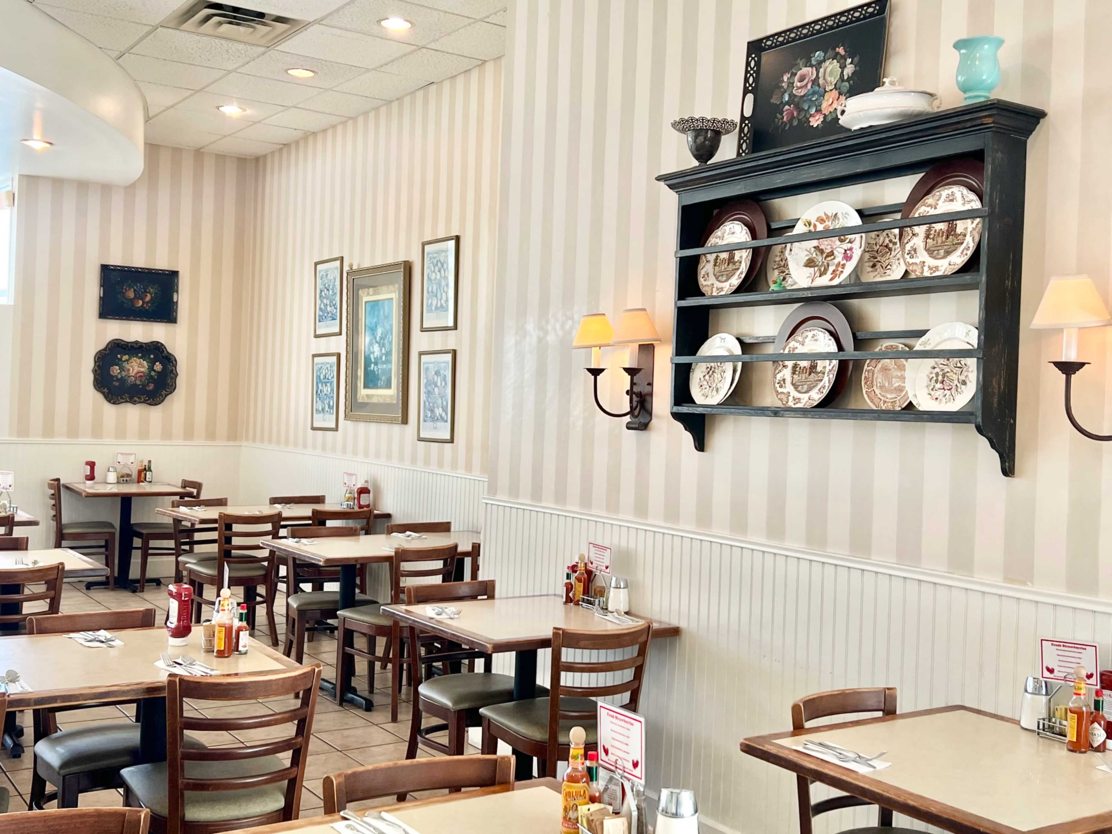 The image shows a cozy restaurant interior with wooden tables, chairs, decorative plates on a shelf, and a light-colored striped wall.