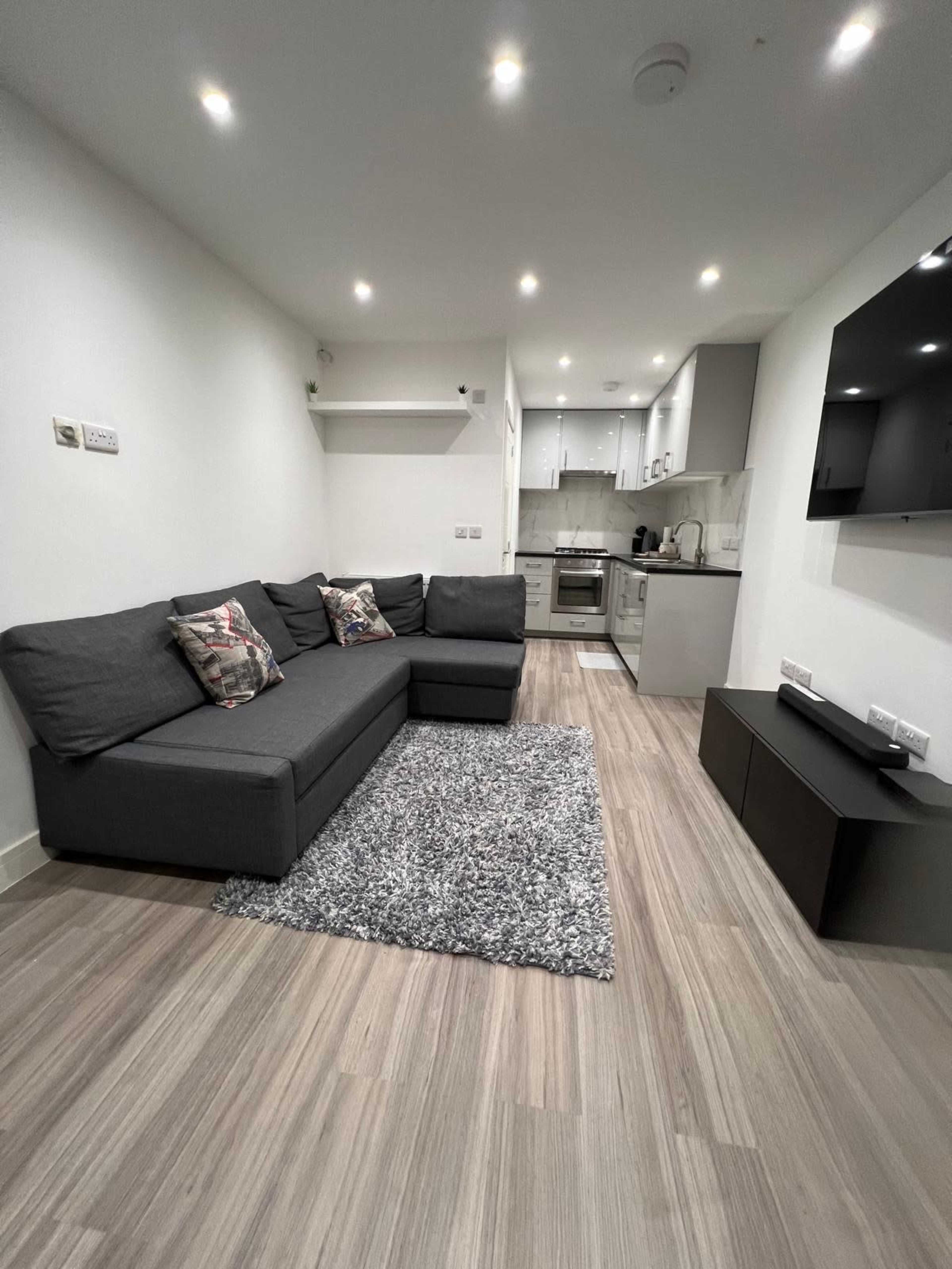 The image shows a modern living room with a gray sectional sofa, a large area rug, and an open kitchen area featuring white cabinetry and stainless steel appliances.