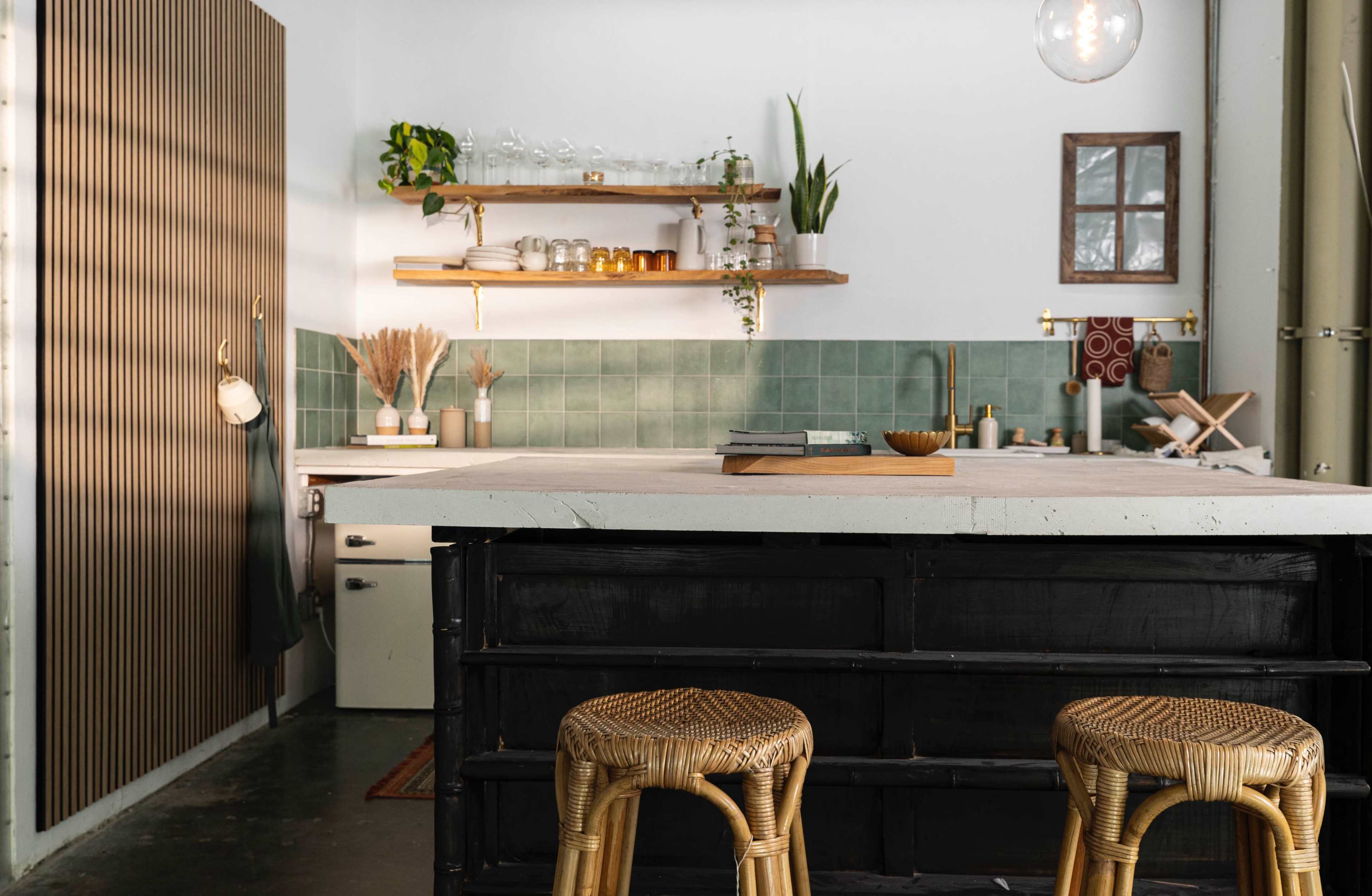 A kitchen features a central island with two woven stools, a green tiled backsplash, wooden shelves with plants and glassware, and a blend of modern and rustic decor elements.