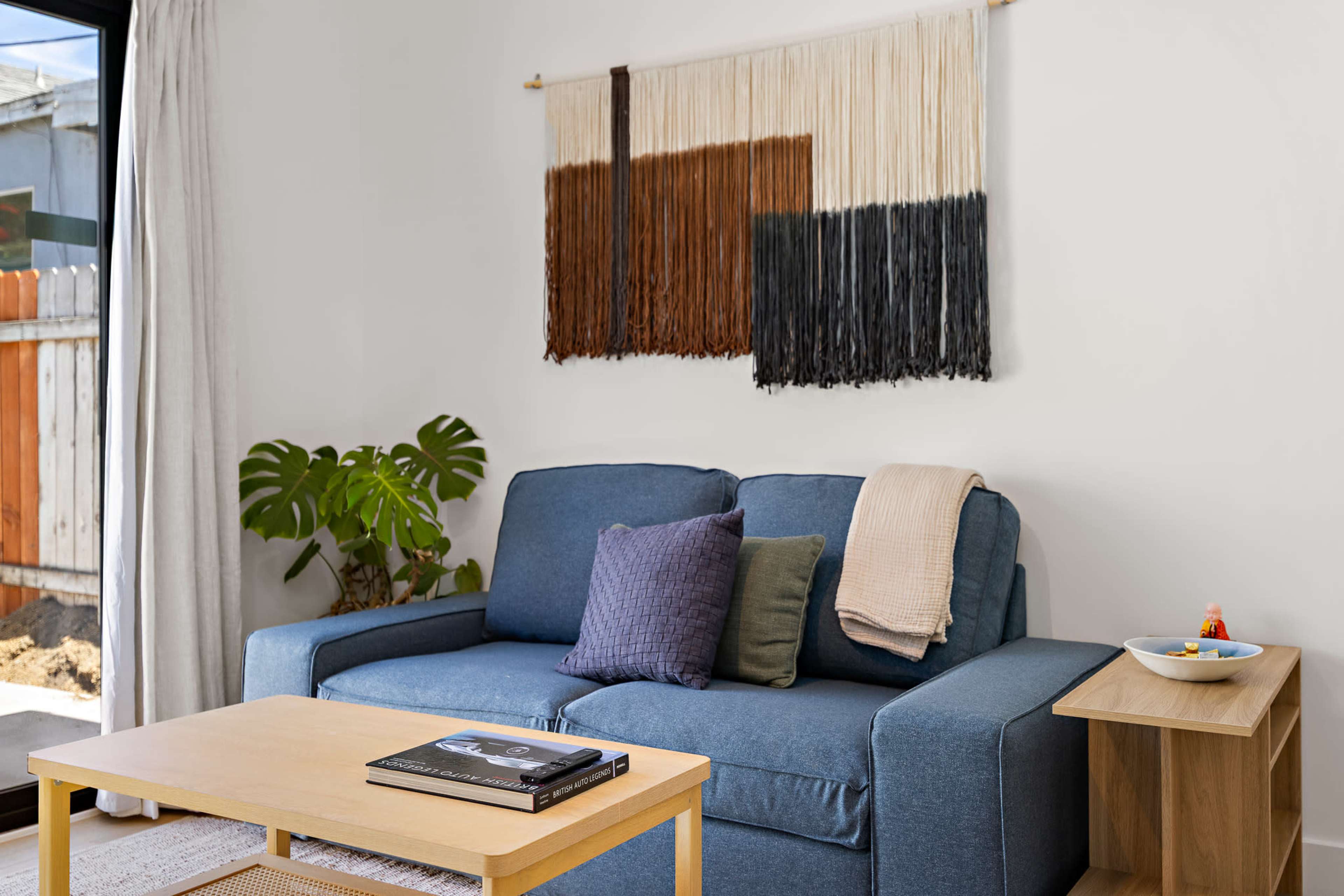 A living room features a blue couch with decorative pillows, a wooden coffee table, and a wall hanging made of yarn in earthy tones.
