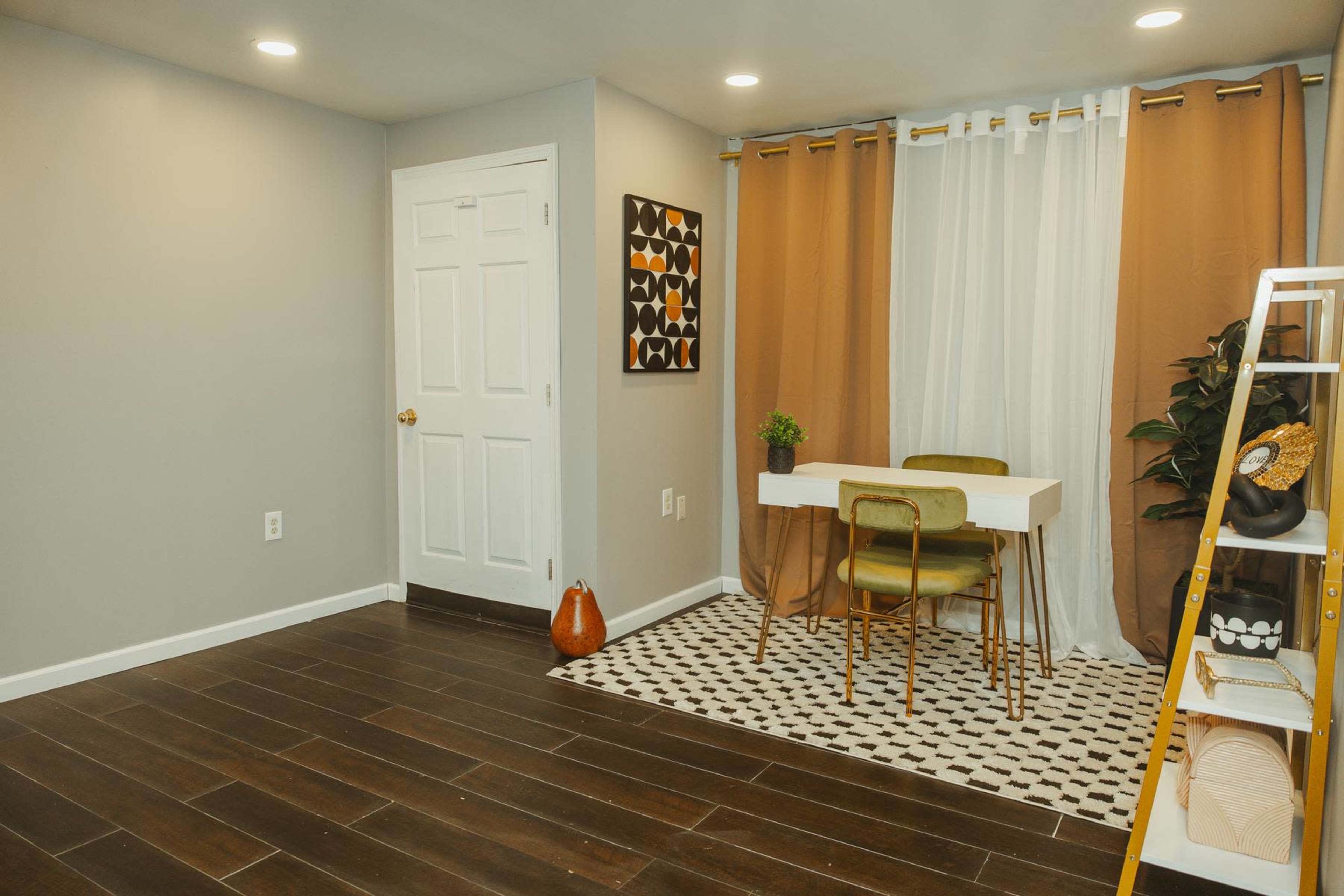 The image shows a small room with a table and chairs, a plant, and a ladder shelf against the wall, featuring light brown and white curtains.