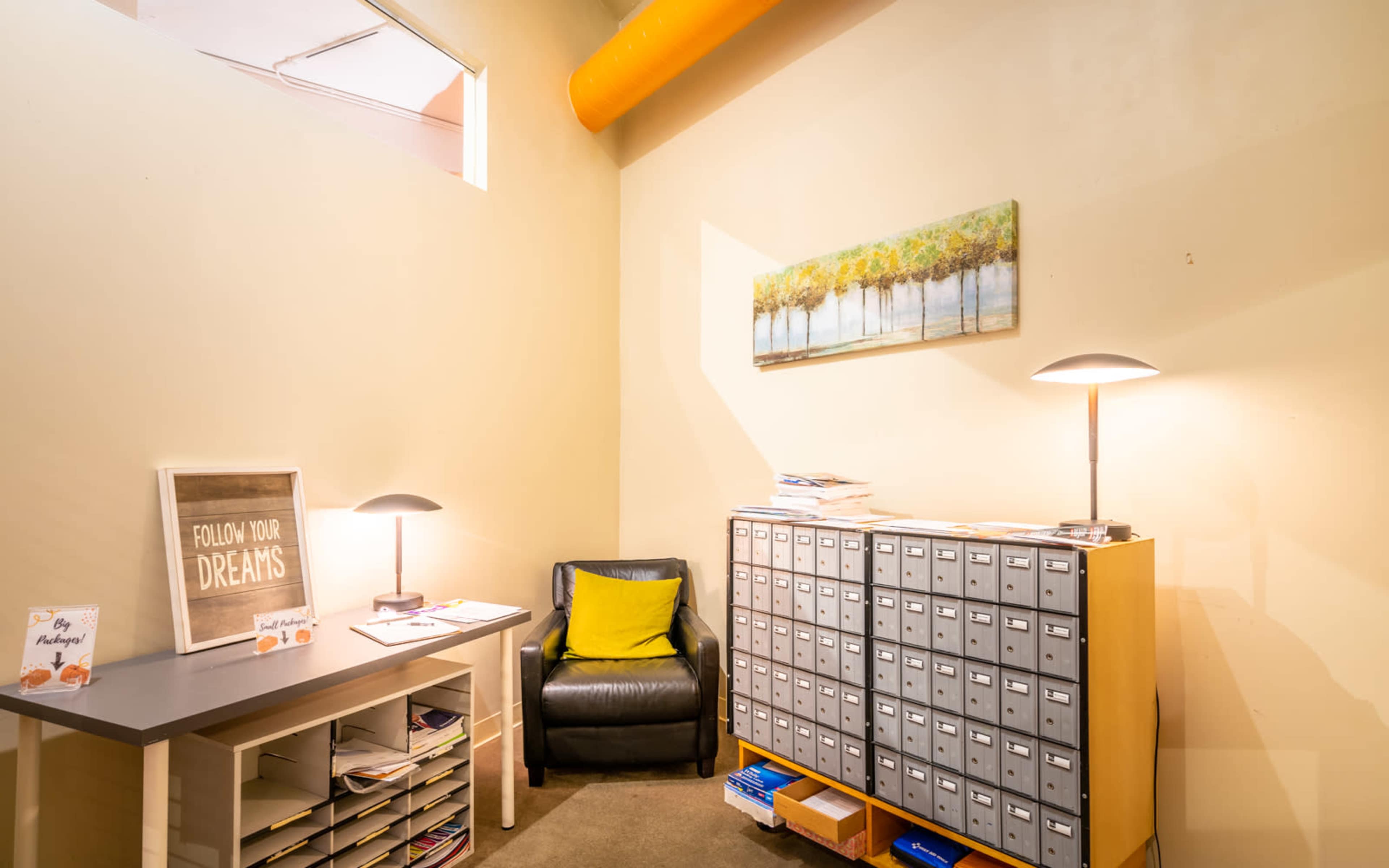 The image shows a corner of a room featuring a black lounge chair, a wooden desk with papers, and a metal mailbox unit against a beige wall.