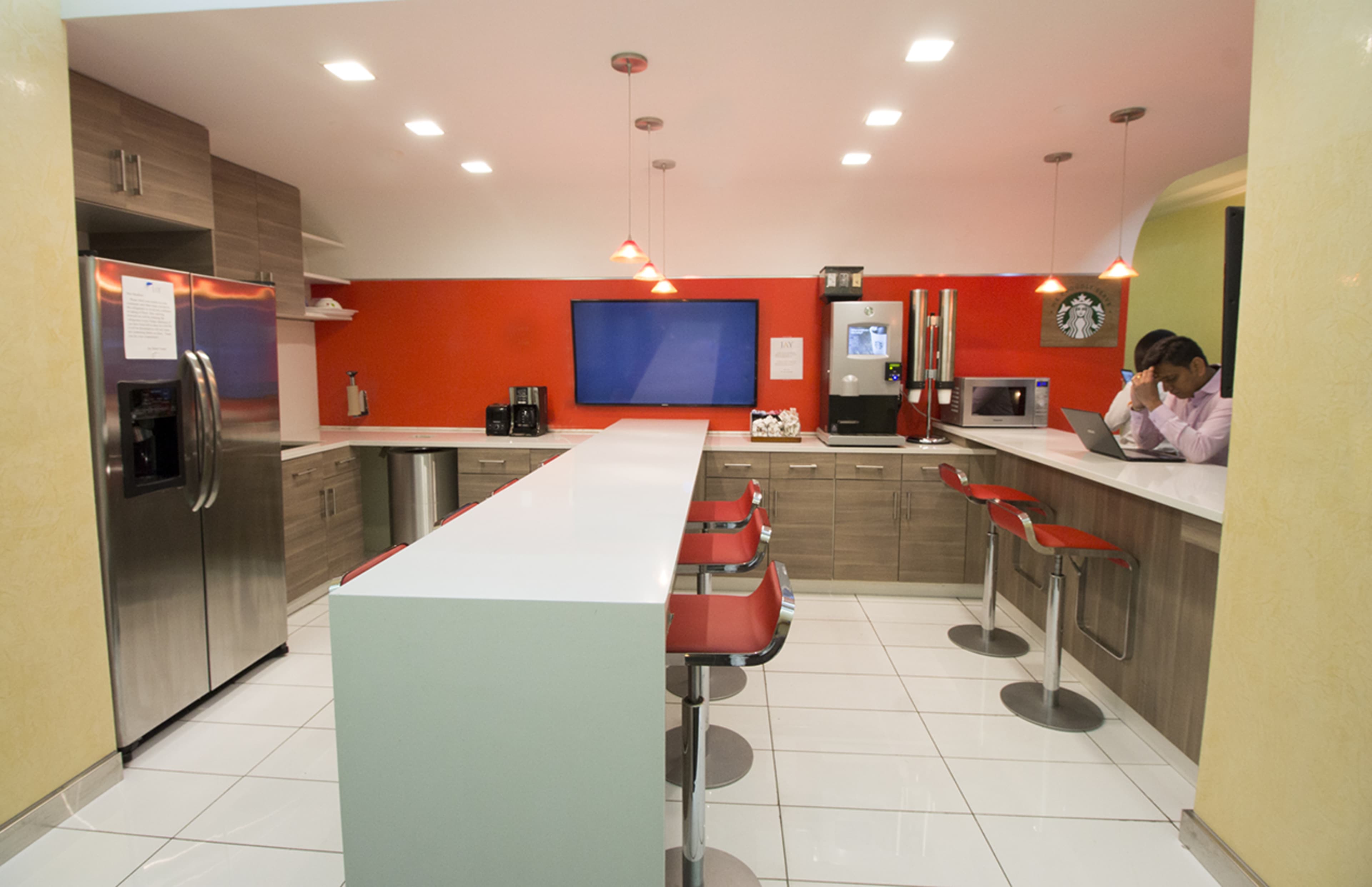 A modern kitchen with stainless steel appliances, a long countertop with red stools, and a man working on a laptop in the background.