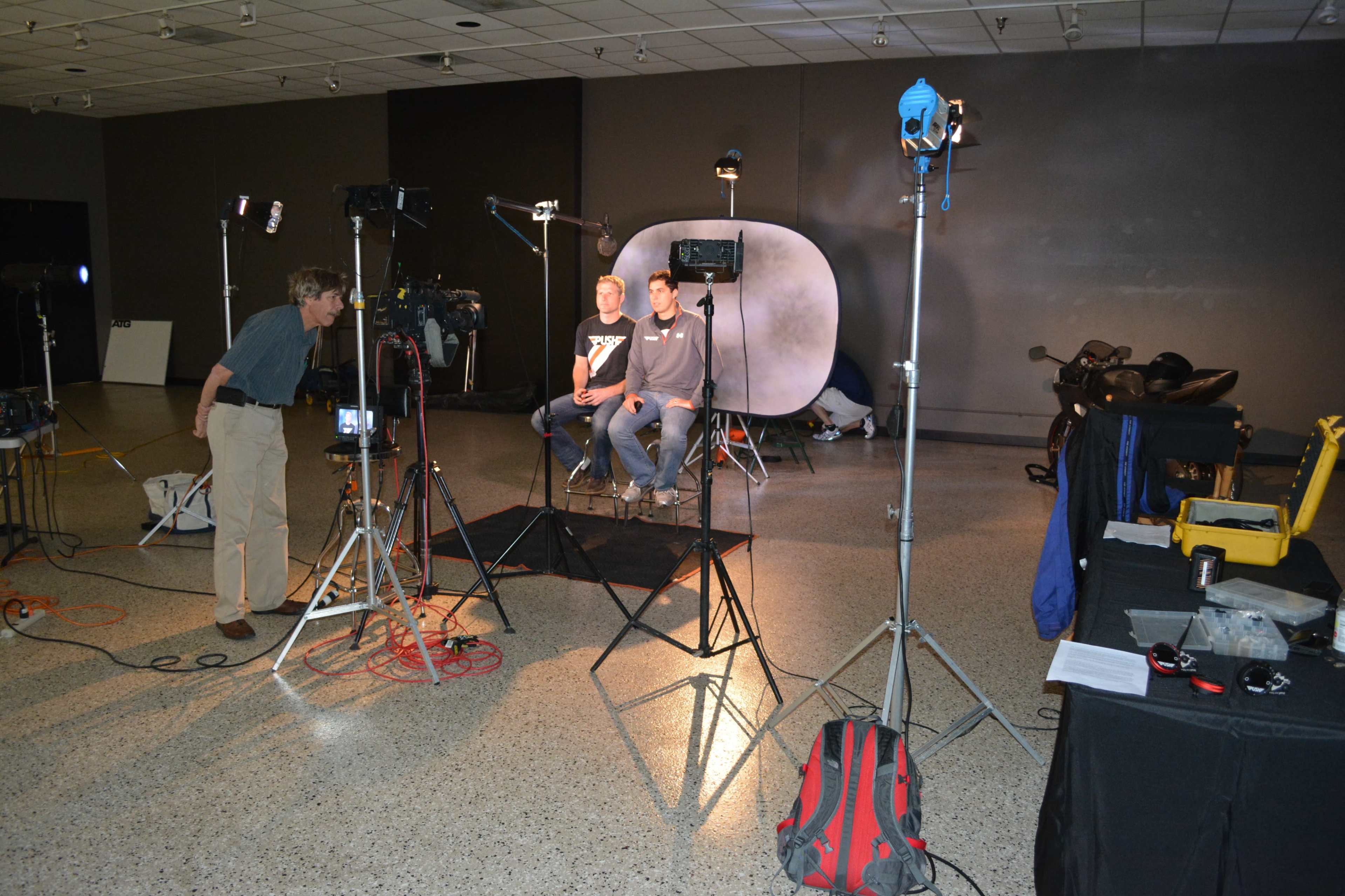 Two individuals sit in a well-lit studio with multiple cameras and lighting setups arranged around them.