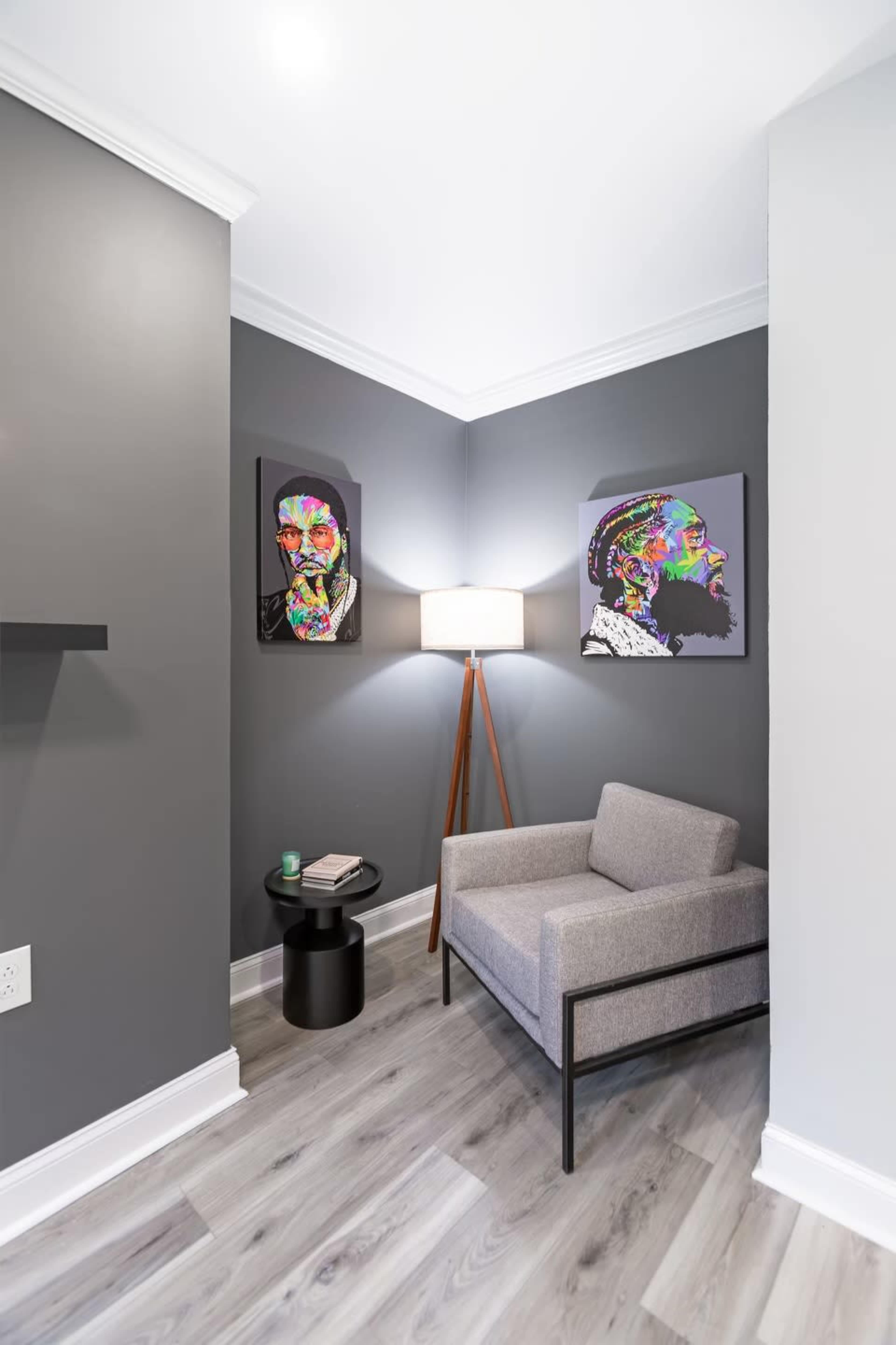 A modern corner seating area features a gray chair beside a small black side table, with vibrant artwork on the walls and a floor lamp illuminating the space.